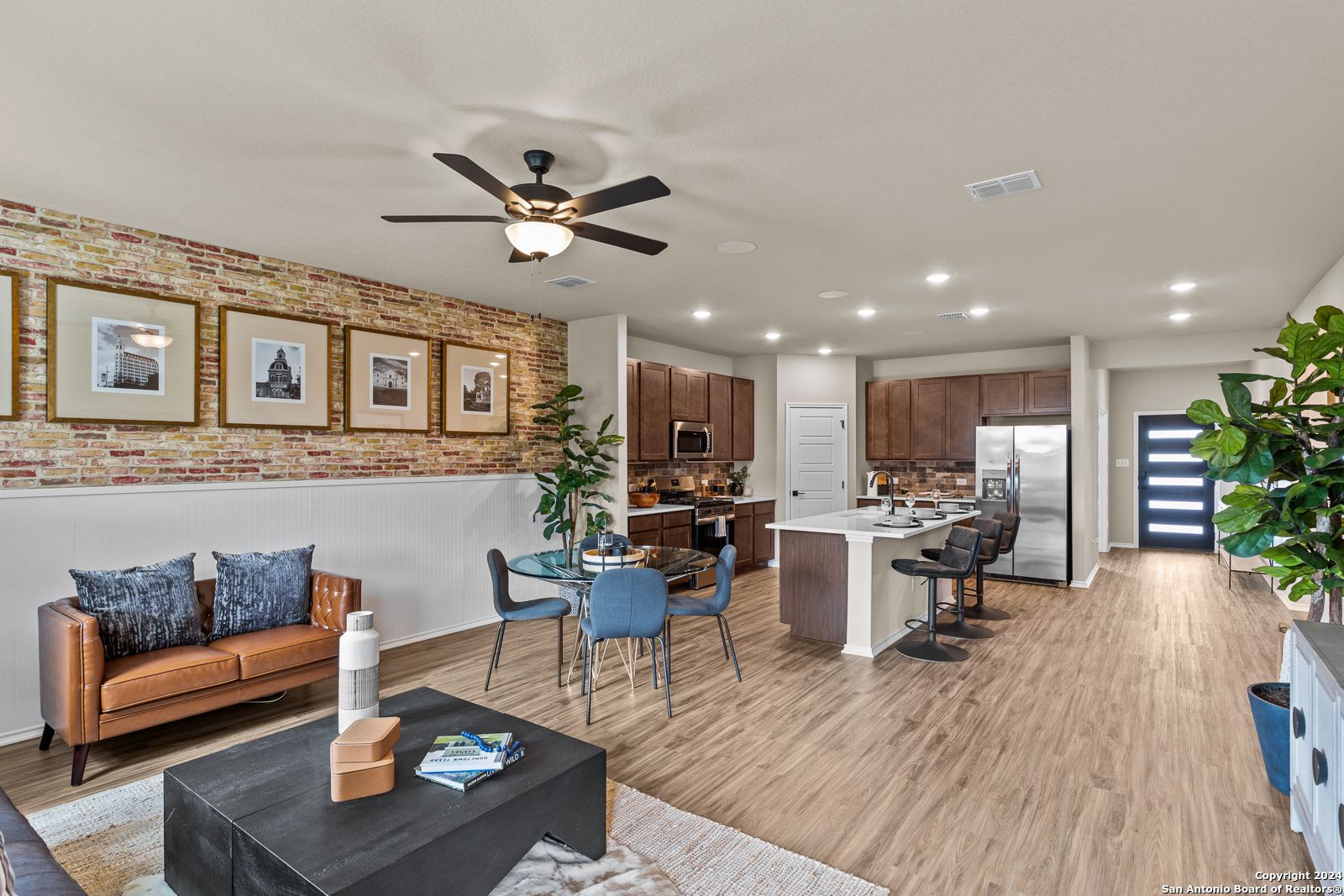 Open-concept living room and kitchen with brick accent wall, leather sofa, island bar, and stainless appliances in Davidson Homes The Douglas F, Bricewood, San Antonio