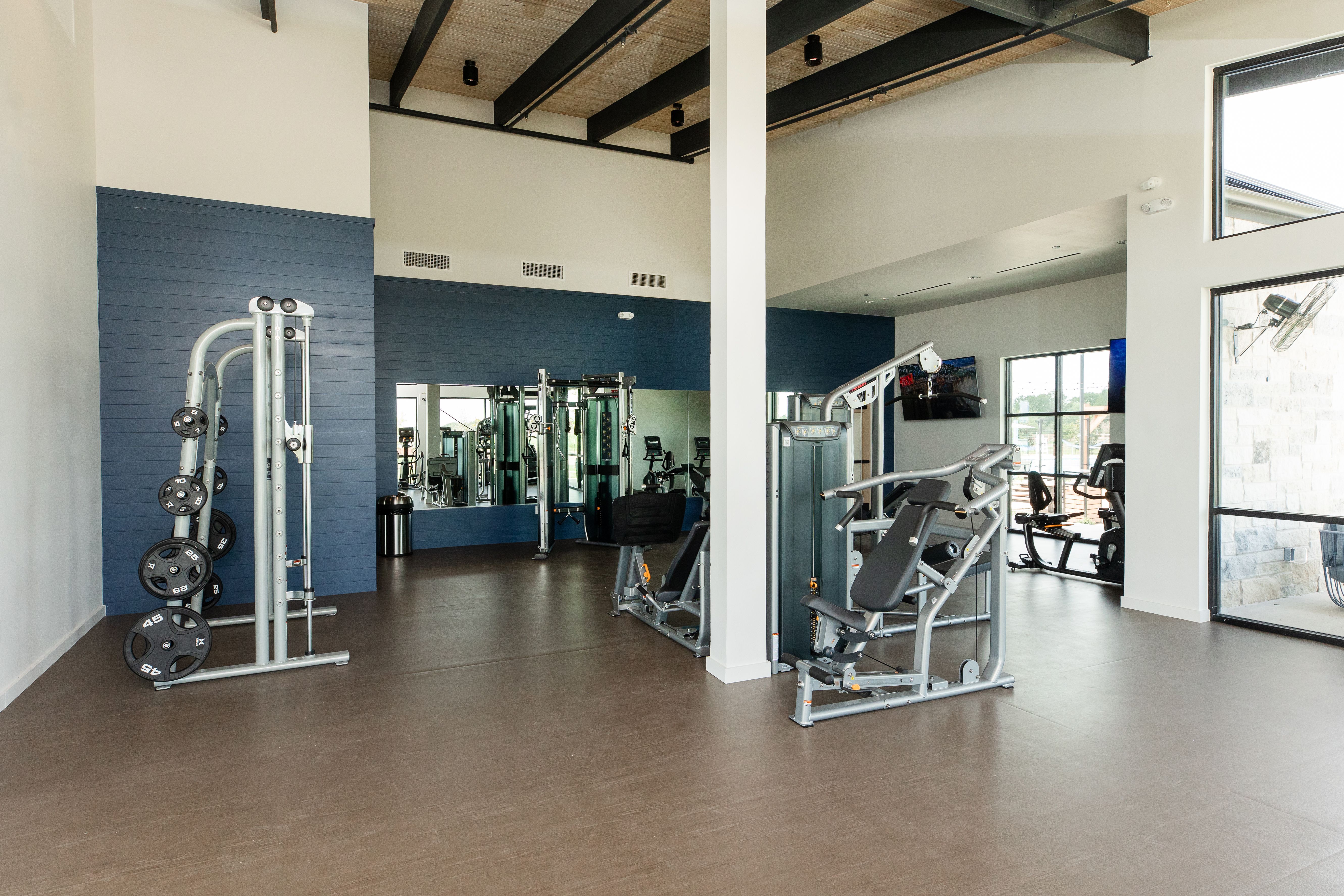 Spacious fitness center at Emberly in Beasley Texas with exposed wooden beams, blue accent wall, mirrors, and weight machines