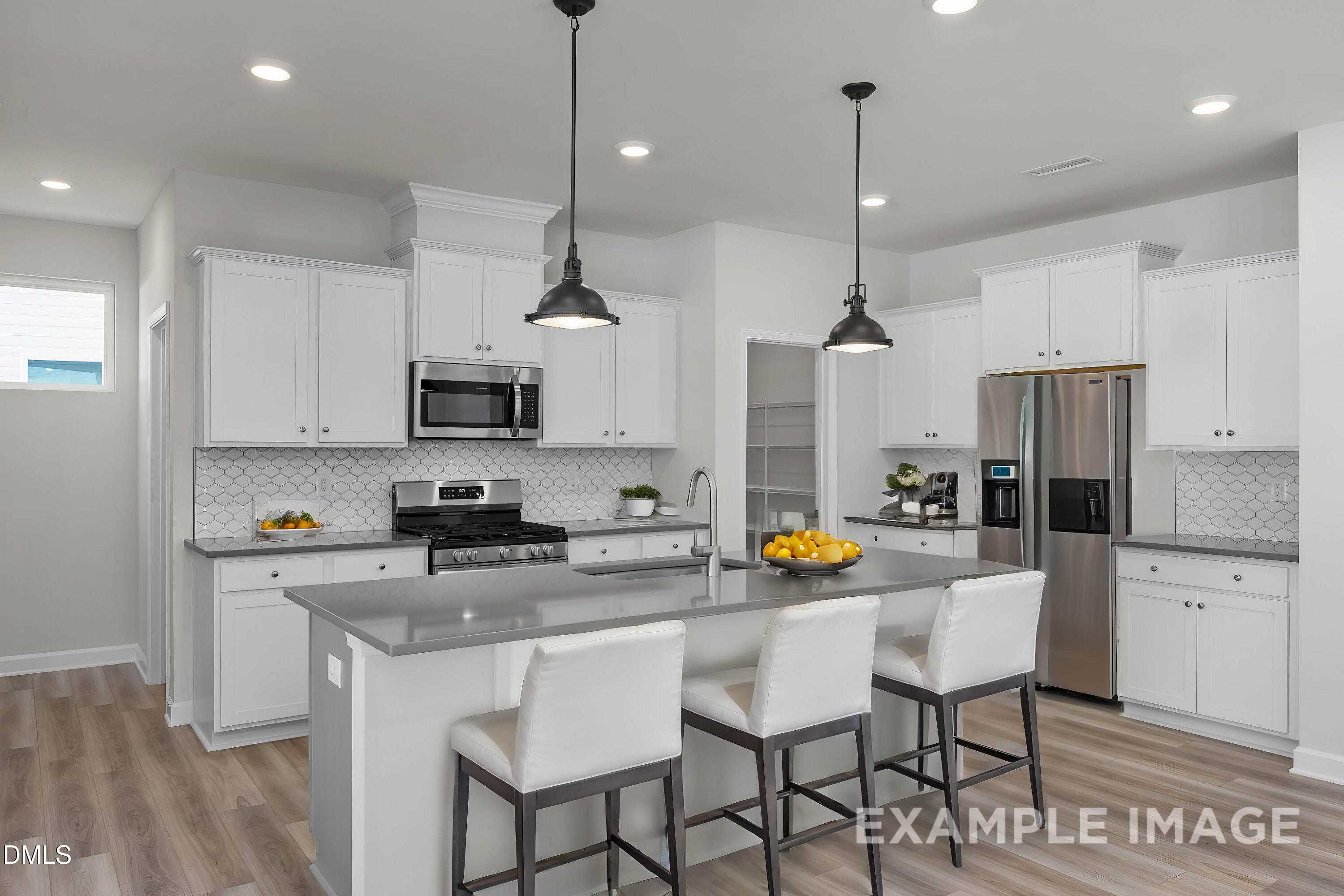 Modern open-concept kitchen with white shaker cabinets, gray quartz counters, stainless appliances, and island bar stools in The Adalynn A, Lillington, NC