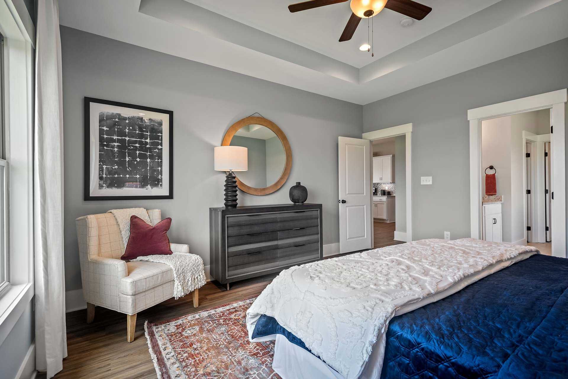 Elegant master bedroom at Heritage Lakes in New Market, Alabama with gray walls, king bed, tufted armchair, and hardwood floors