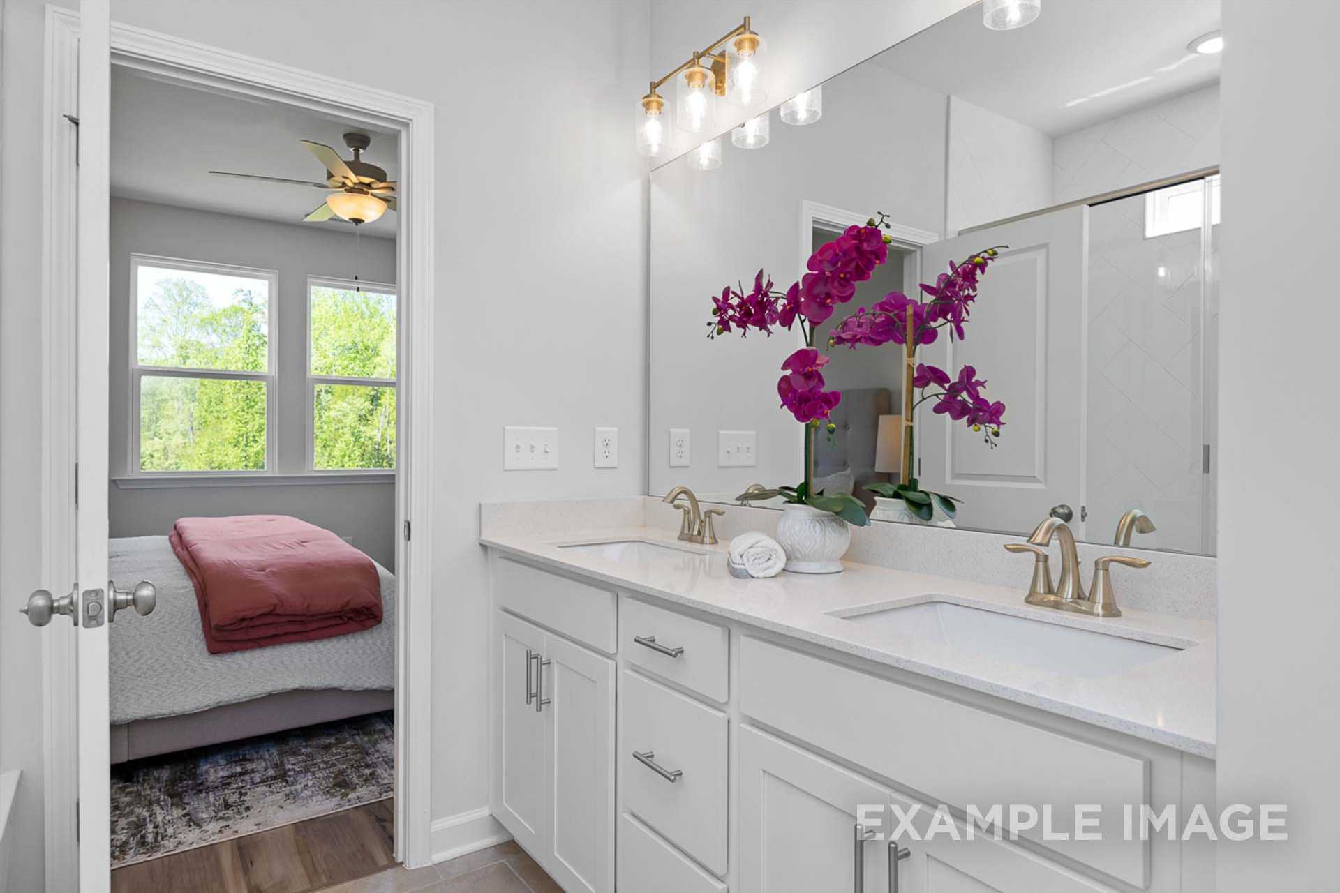 Master bathroom in The Carter B showcasing double vanity with gold faucets, purple orchids, and open bedroom view