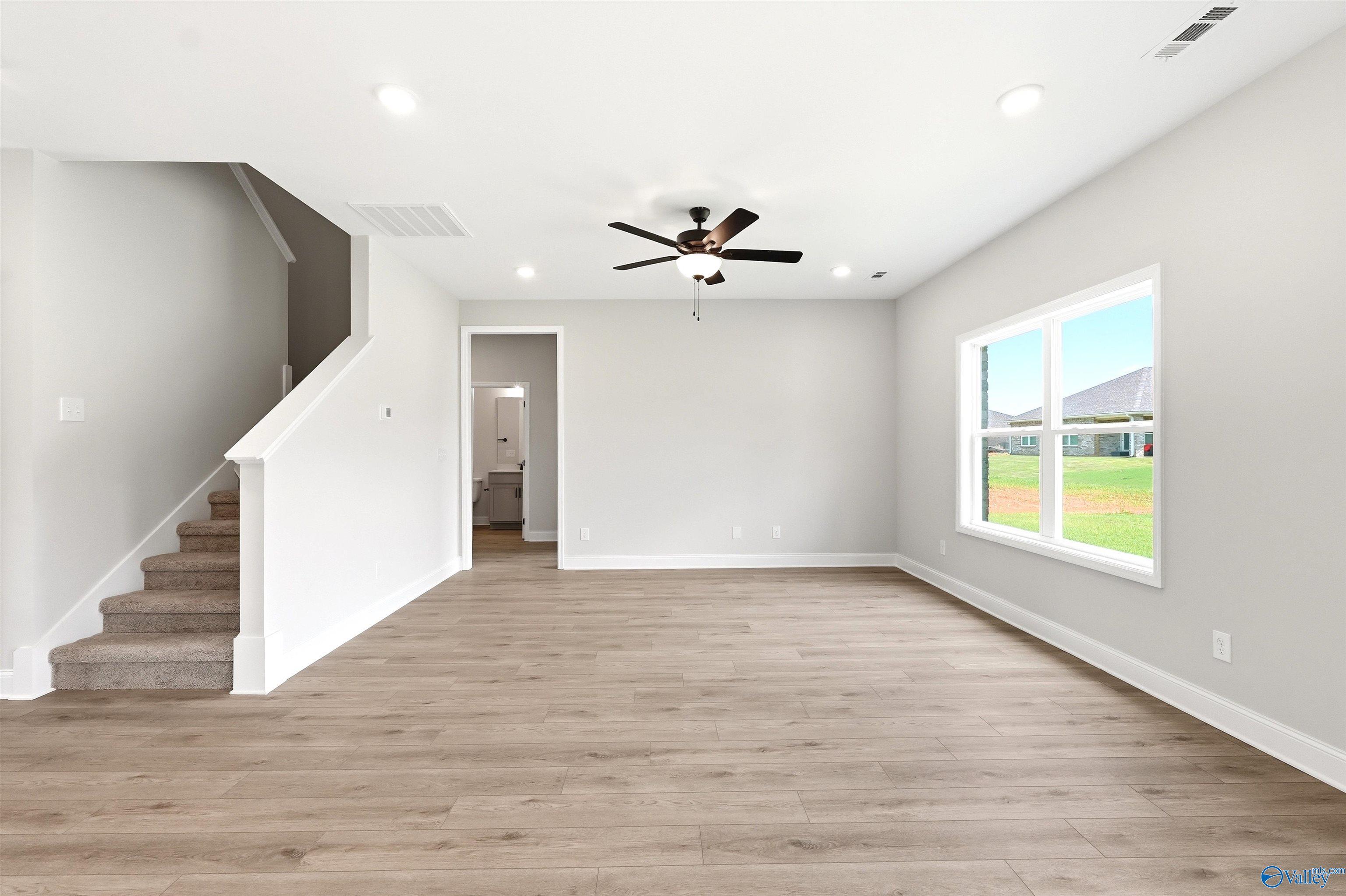 Spacious open living room with light wood floors, ceiling fan, and carpeted stairs in The Shelby A home at Wood Trail.