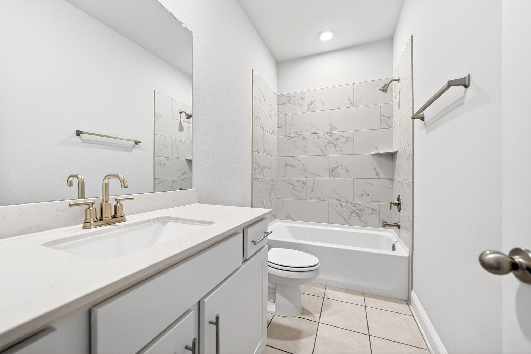 Modern bathroom in The Redbud featuring a marble-style tiled tub-shower combo, white vanity, and elegant gold fixtures.