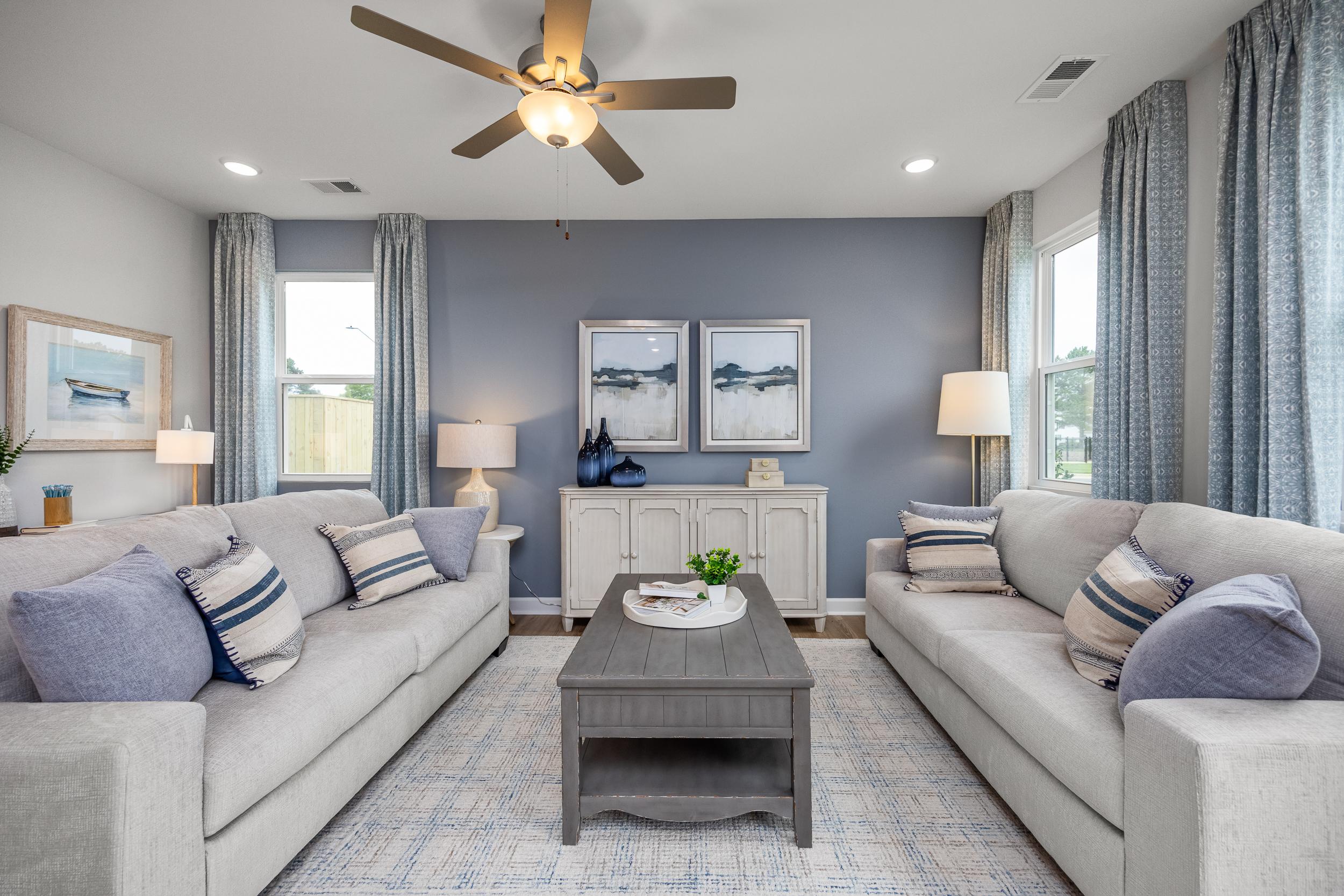 Spacious living room at Springvale in Fuquay-Varina NC with light gray sofas, blue accent wall, ceiling fan, and coastal artwork