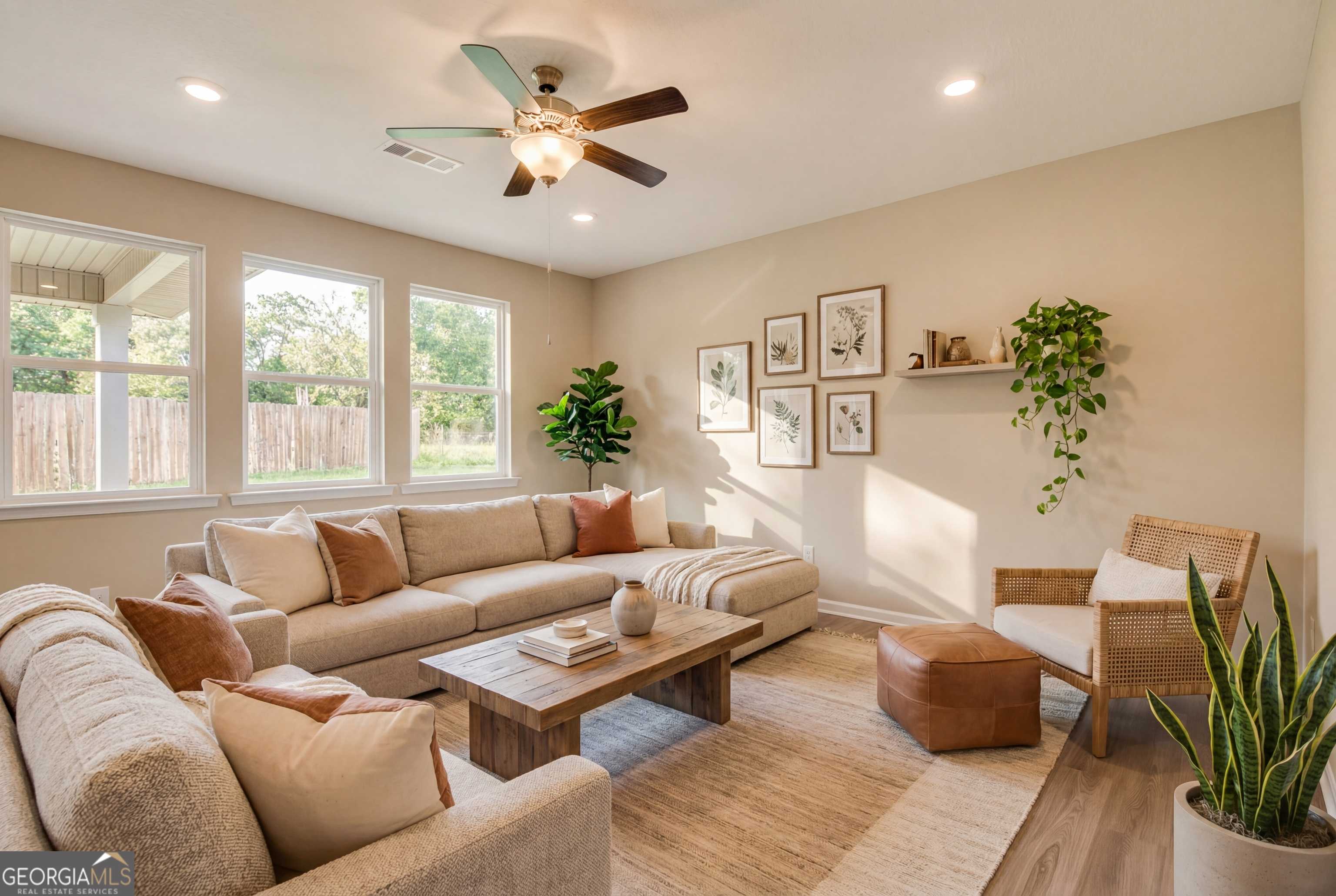 Cozy living room with beige sectional sofa, wooden coffee table, rattan chair, potted plants, and large windows in Evermore Homes The Luna, Ivy Glen, Perry, Georgia