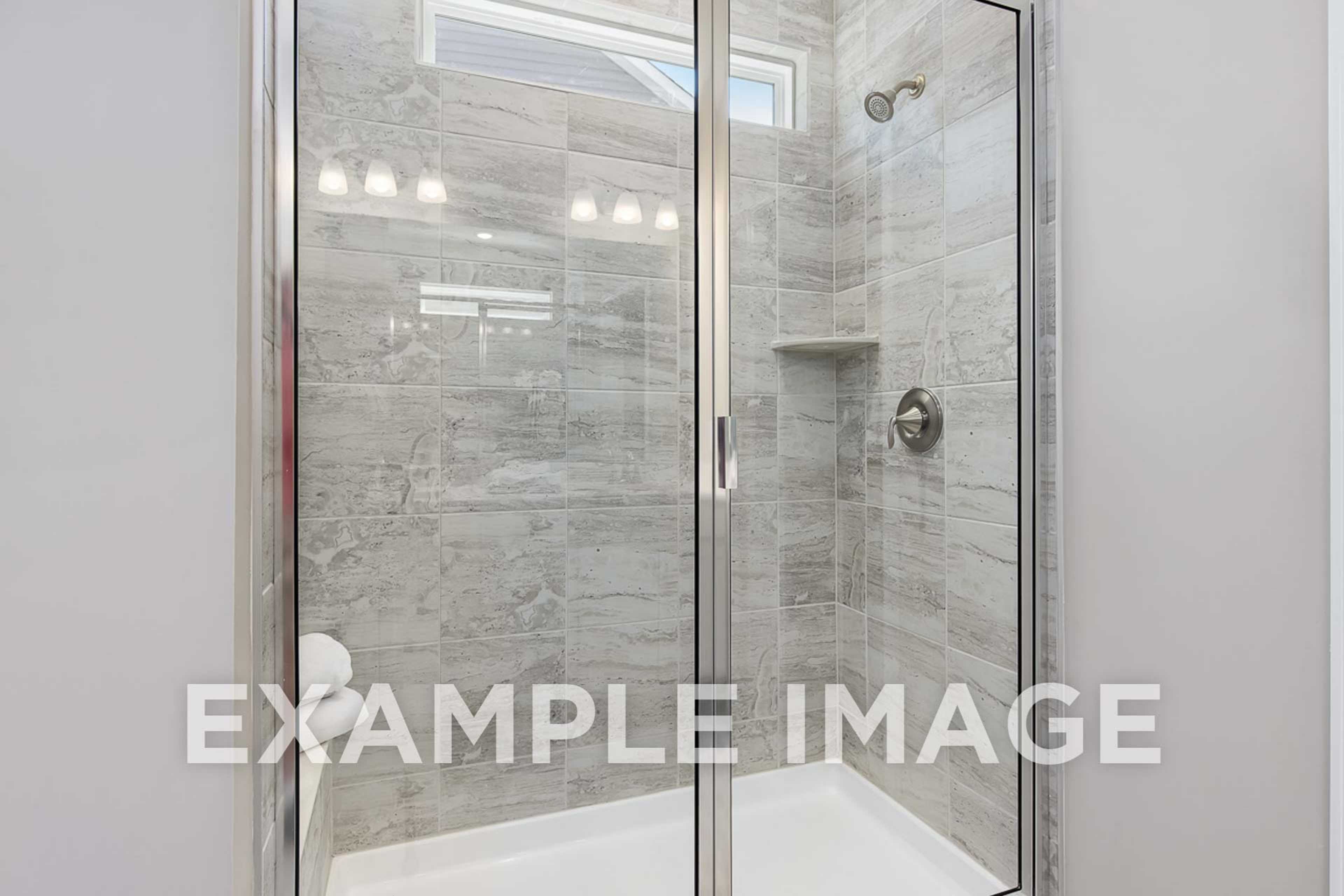 Spacious frameless glass shower in The Ash D master bathroom with gray tile walls, rainfall showerhead, and built-in niche