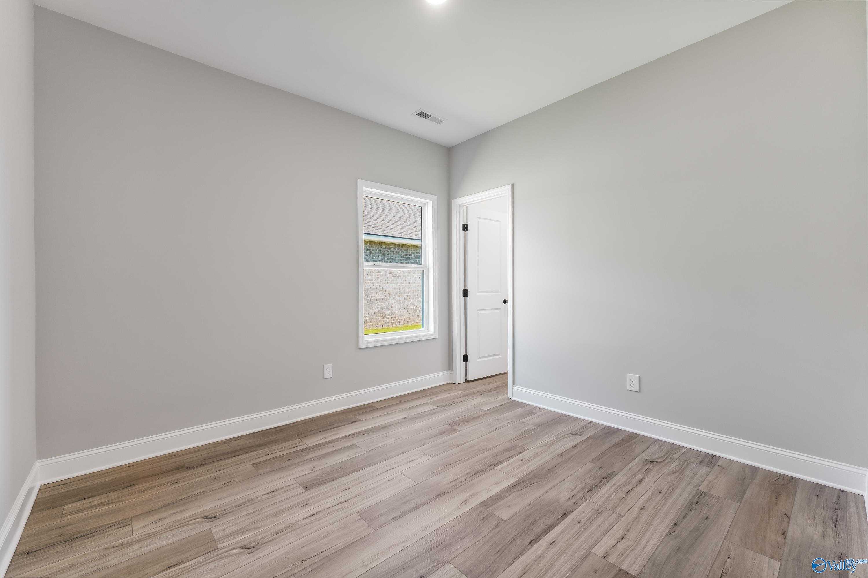 Empty bedroom with gray walls, window blinds, white door, and light hardwood floors in Davidson Homes The Daphne C, Hazel Green, AL