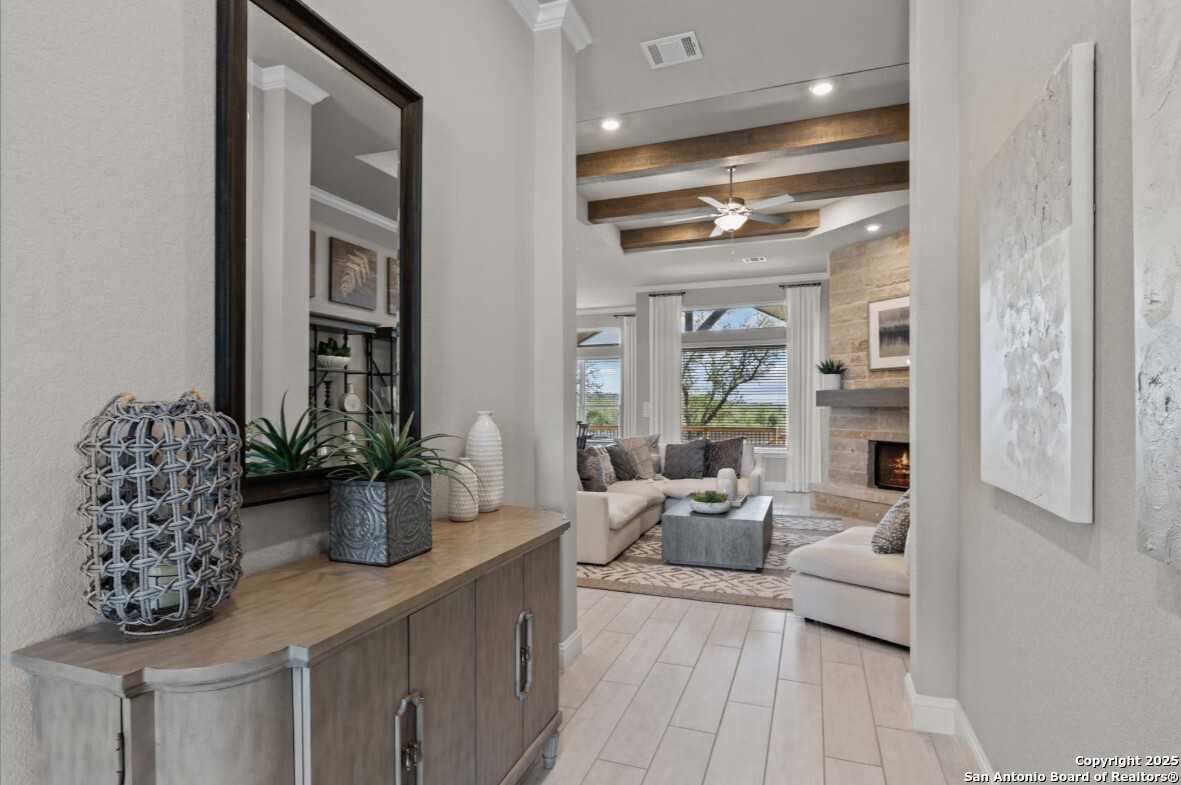 Elegant entryway with mirrored console and plants opening to living room featuring stone fireplace, beige sofa, and beamed ceiling in Davidson Homes The Garner B, Ladera, San Antonio