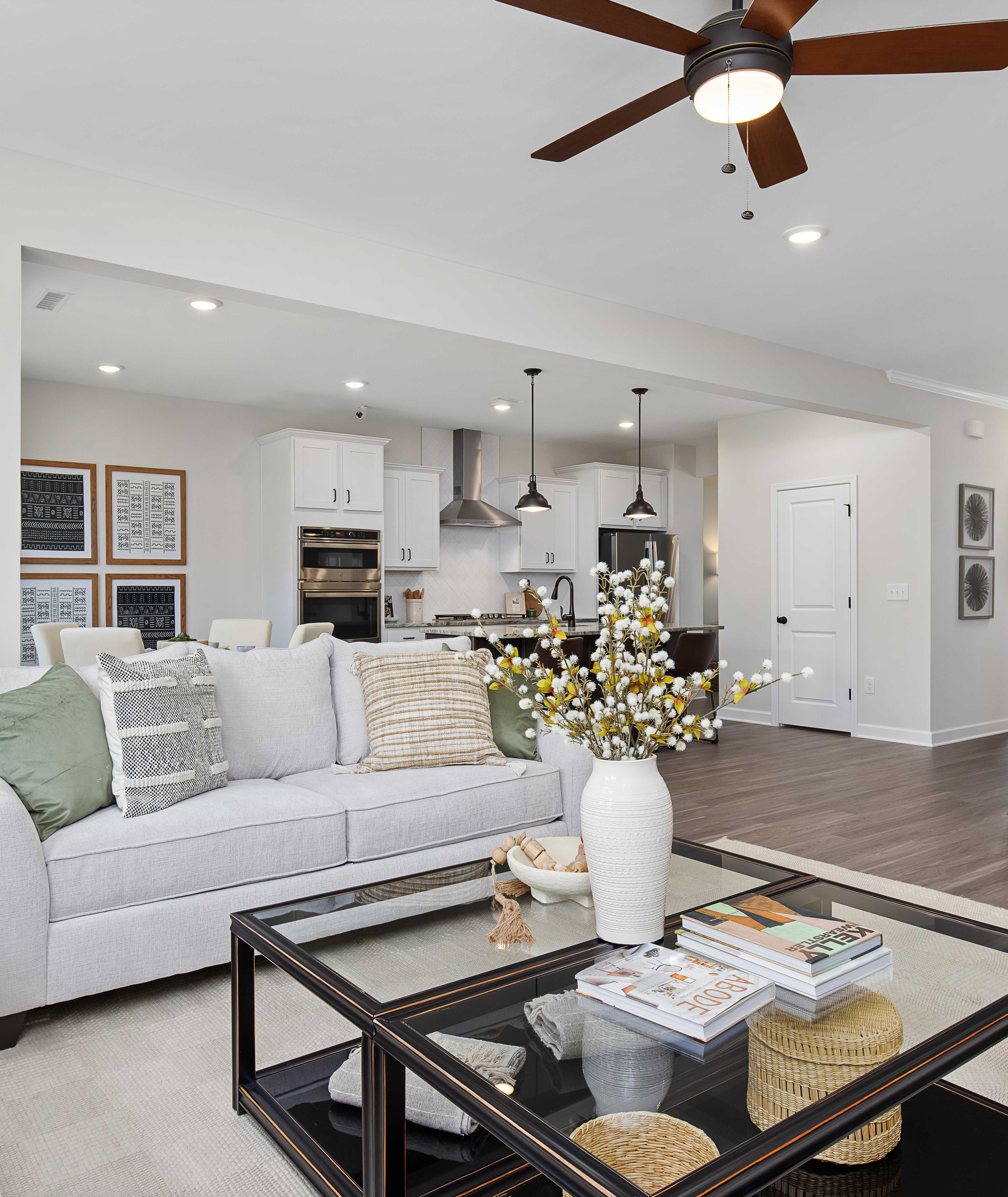 Spacious modern living room with white walls, open white kitchen, light gray sofa with green pillows, glass coffee table, har