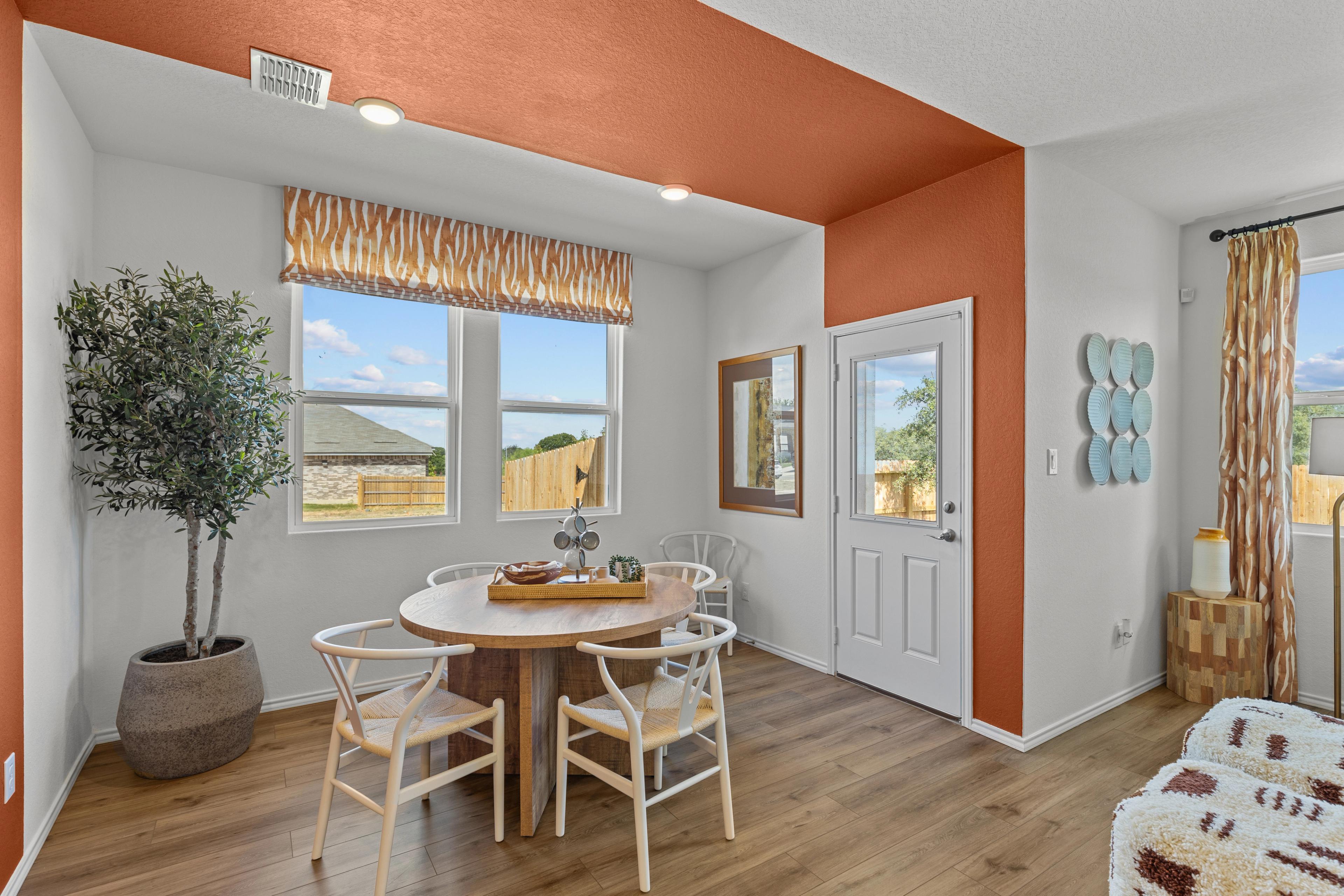 Cozy dining nook in The Daphne G home with round wooden table, orange accent wall, potted olive tree, and backyard views