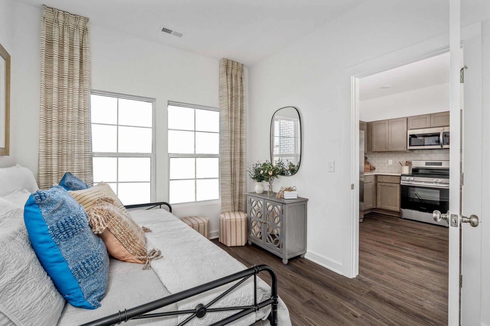 Cozy bedroom nook with metal daybed, blue pillows, sheer curtains on windows, round mirror, and open kitchen view in Davidson Homes Cumberland A, Gallatin TN