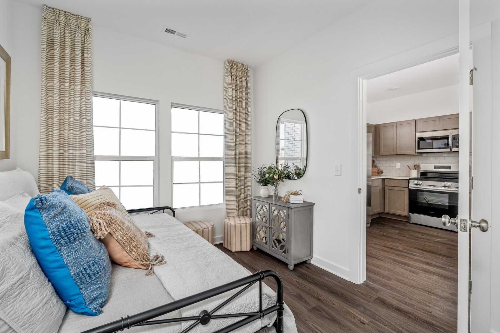 Cozy bedroom nook with metal daybed, blue pillows, sheer curtains on windows, round mirror, and open kitchen view in Davidson Homes Cumberland A, Gallatin TN