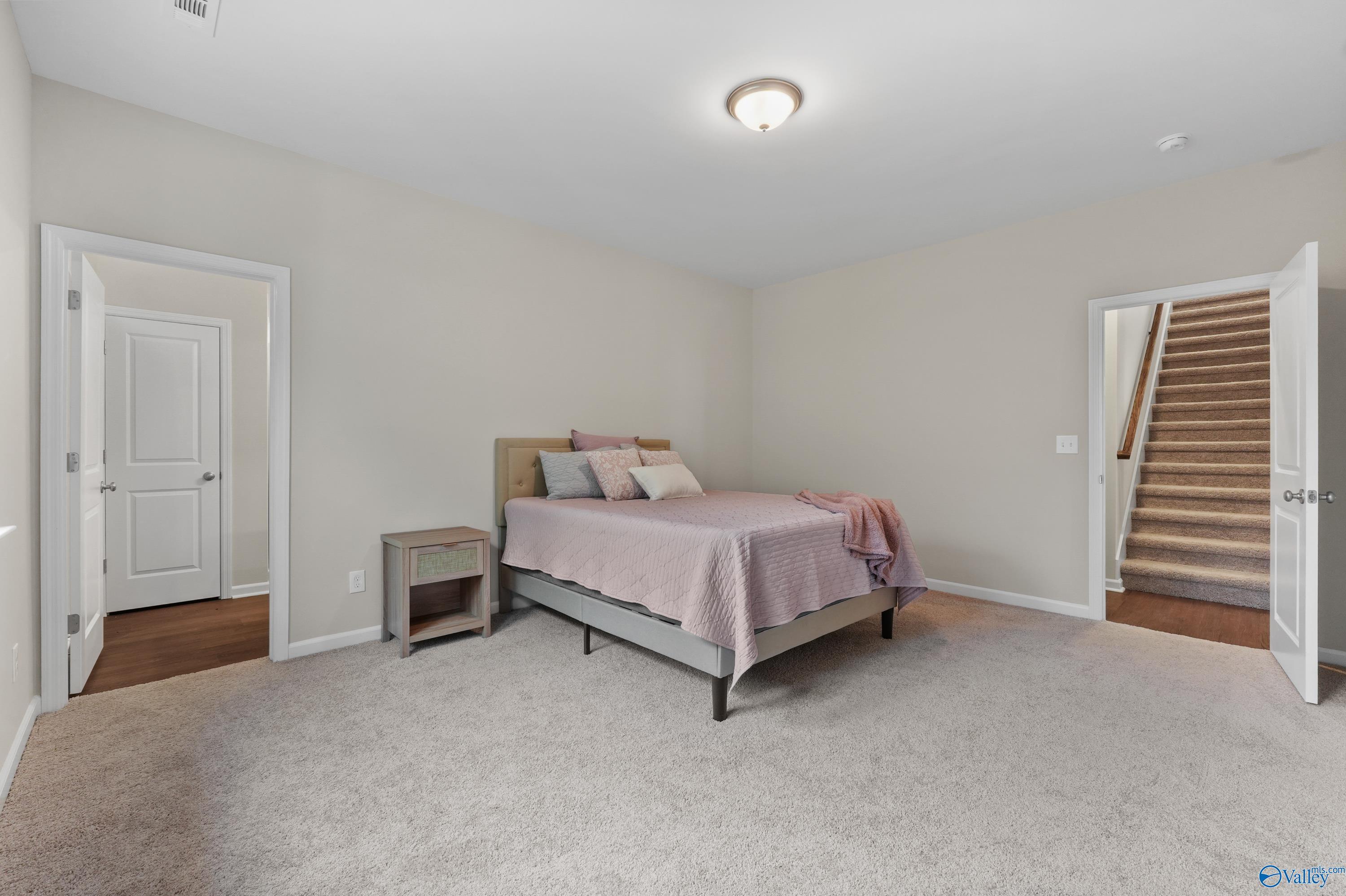 Cozy secondary bedroom with queen bed, pink bedding, nightstand, hallway access, and stairs in Davidson Homes The Stella, Hazel Green, Alabama