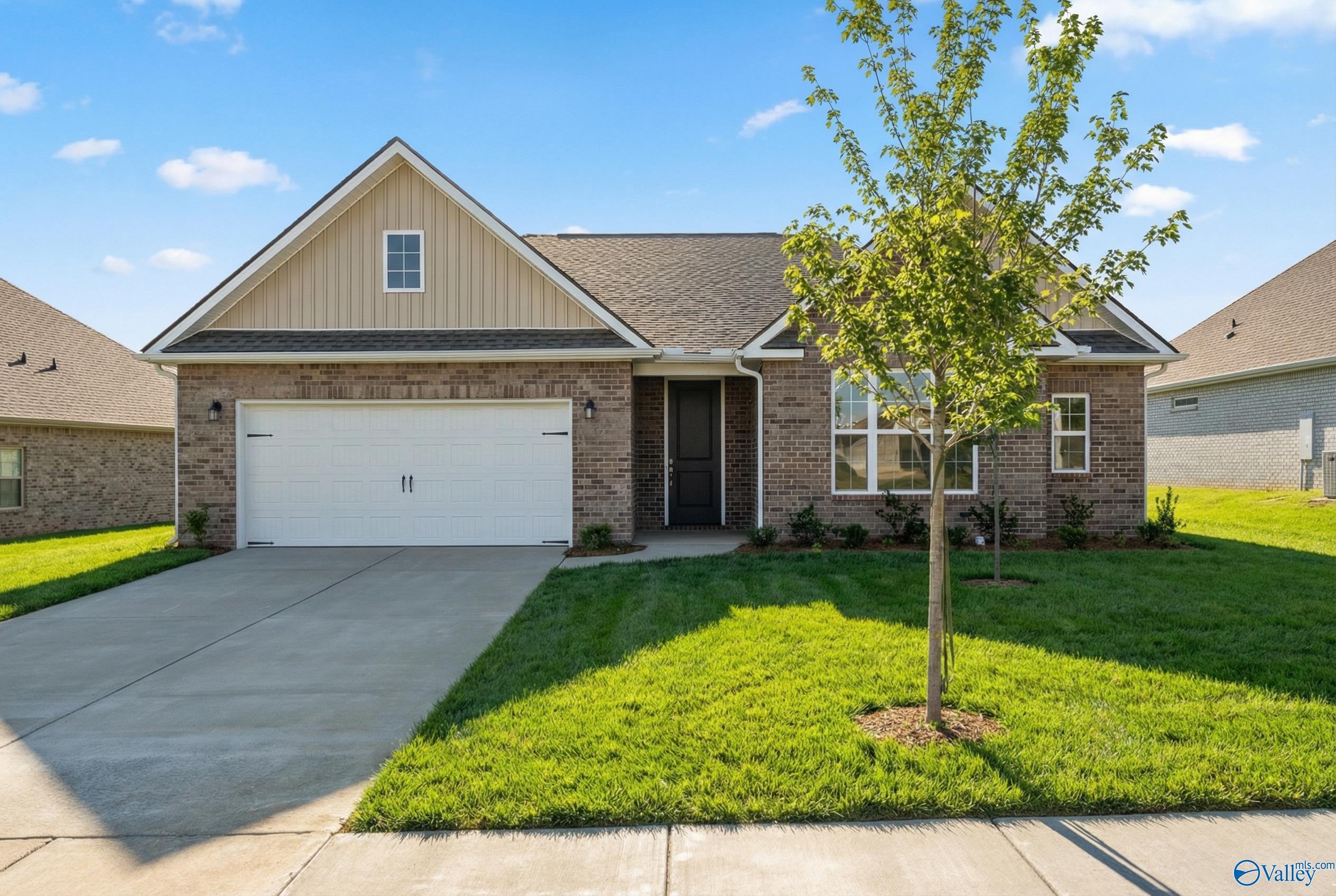 Beige brick 1-story home with 2-car garage, gabled roof, manicured lawn, and young tree in Creekside, Harvest, Alabama by Davidson Homes