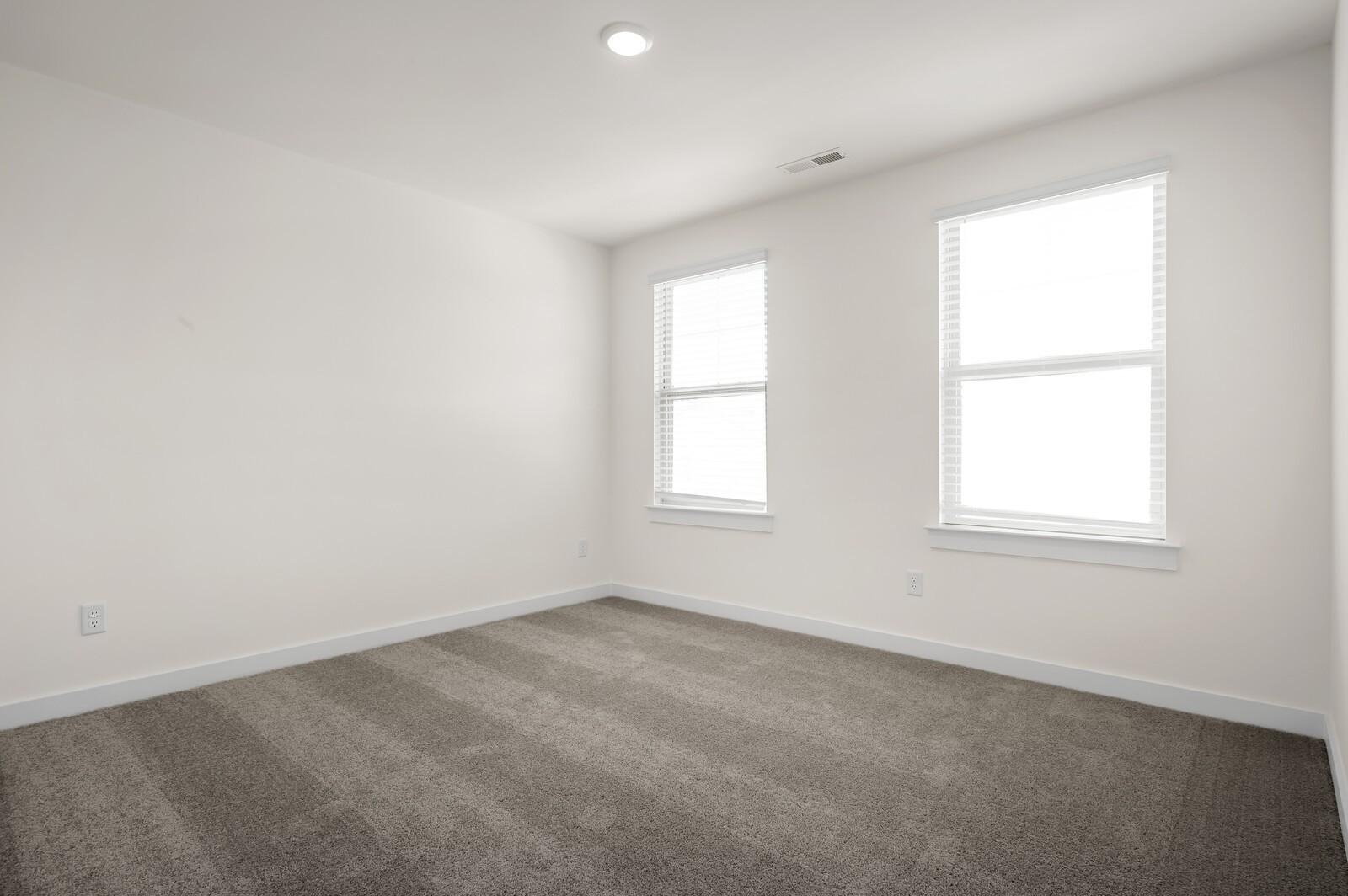 Bright, carpeted bedroom with neutral walls and two large windows in The Ash A floor plan at Woods Crossing, Gallatin.