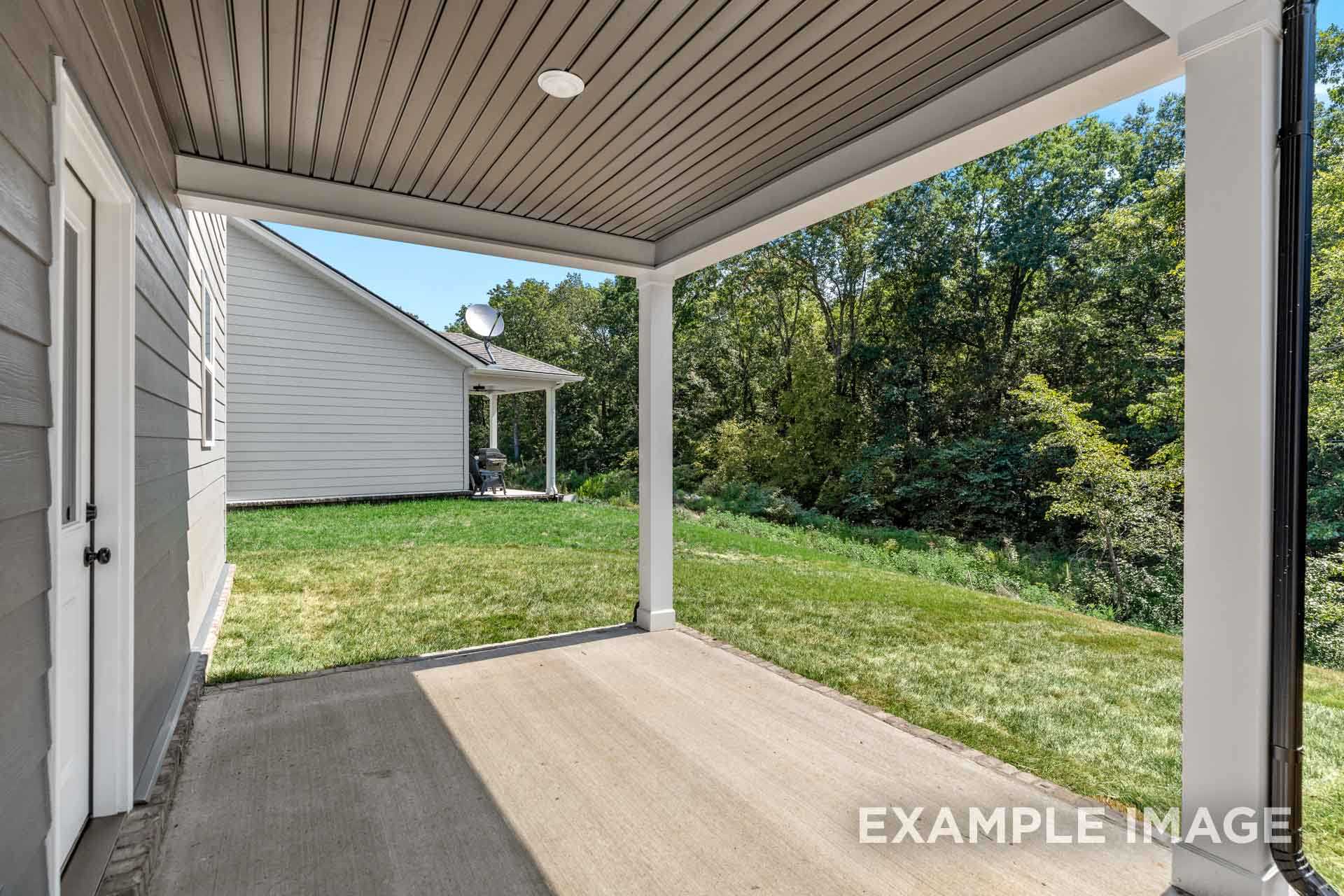 Covered back porch of The Henry B home featuring gray siding, white columns, lush green yard, and wooded view in Mt. Juliet