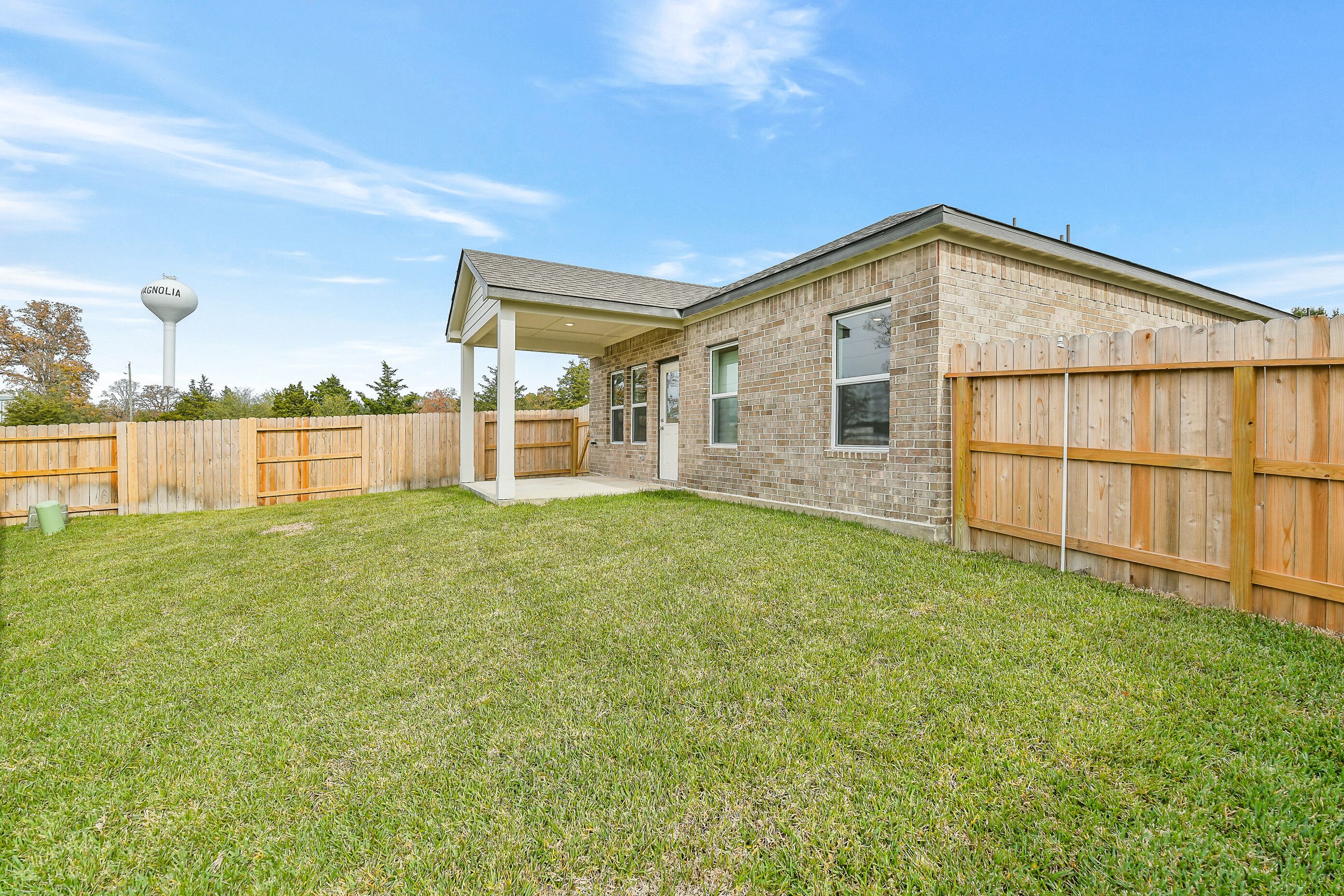 Single-story brick home with covered porch, wooden fence, lush green lawn, and Magnolia water tower backdrop in Windmill Estates, Texas