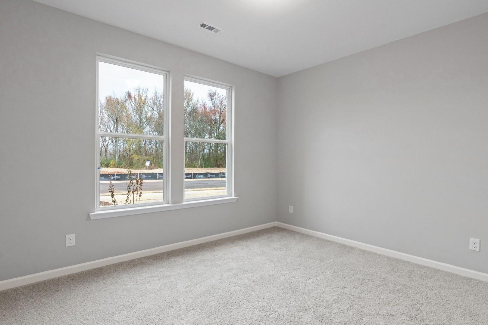 Spacious empty bedroom in The Luna home with gray walls, large double windows overlooking trees, and neutral carpet flooring
