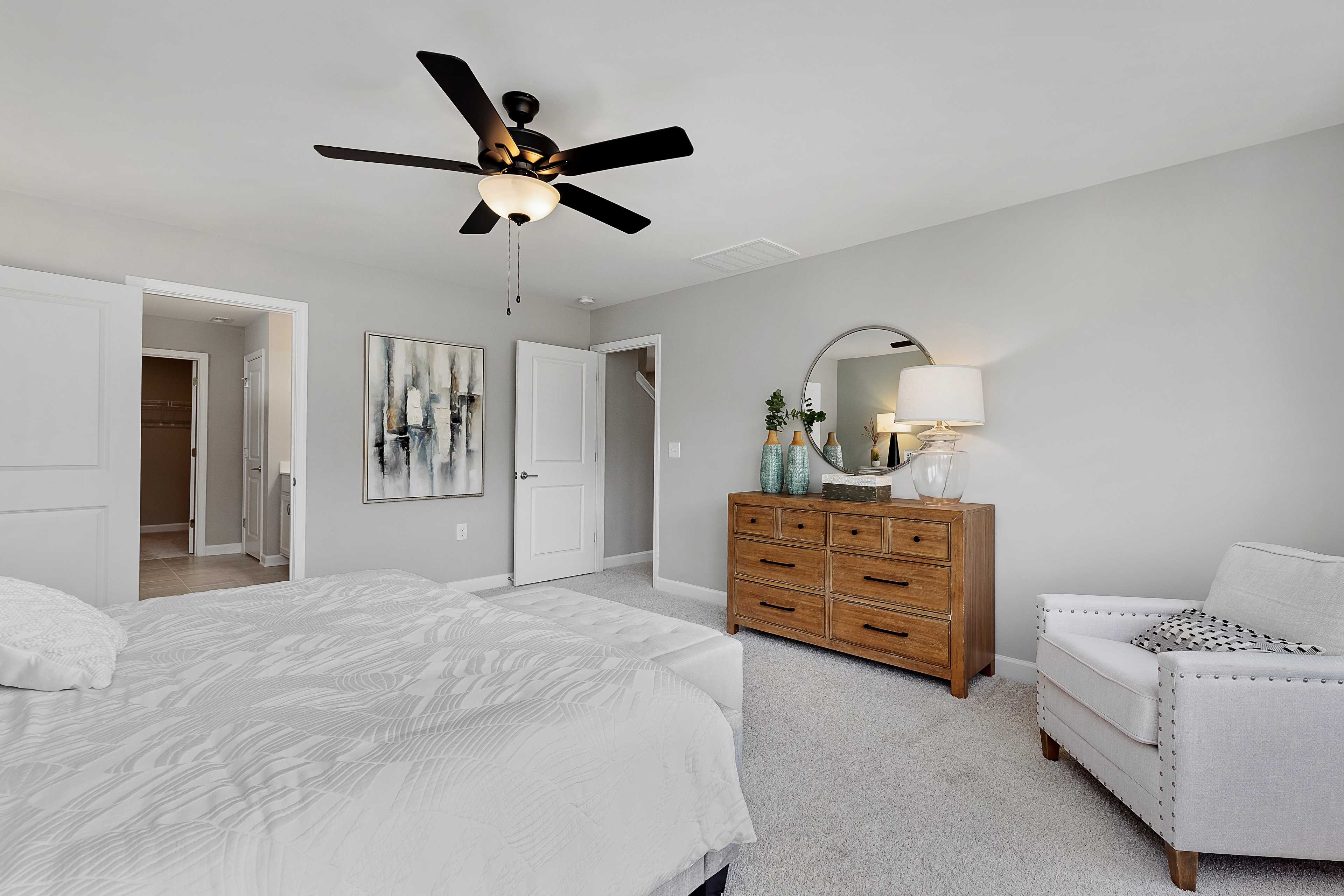 Spacious primary bedroom at Woodland Crossing in Zebulon NC by Davidson Homes with king bed, oak dresser, round mirror, ceiling fan