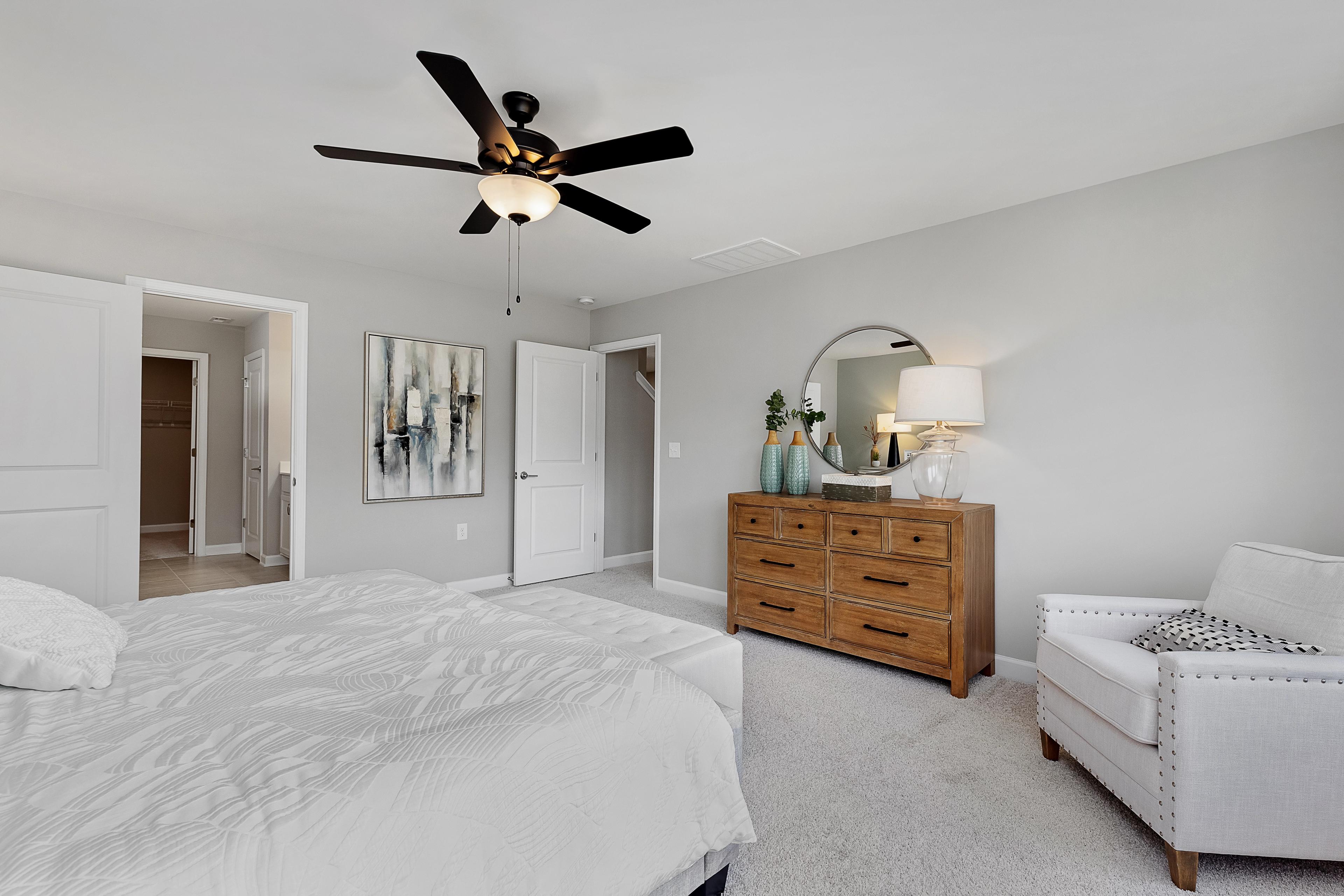Spacious primary bedroom at Woodland Crossing in Zebulon NC by Davidson Homes with king bed, oak dresser, round mirror, ceiling fan