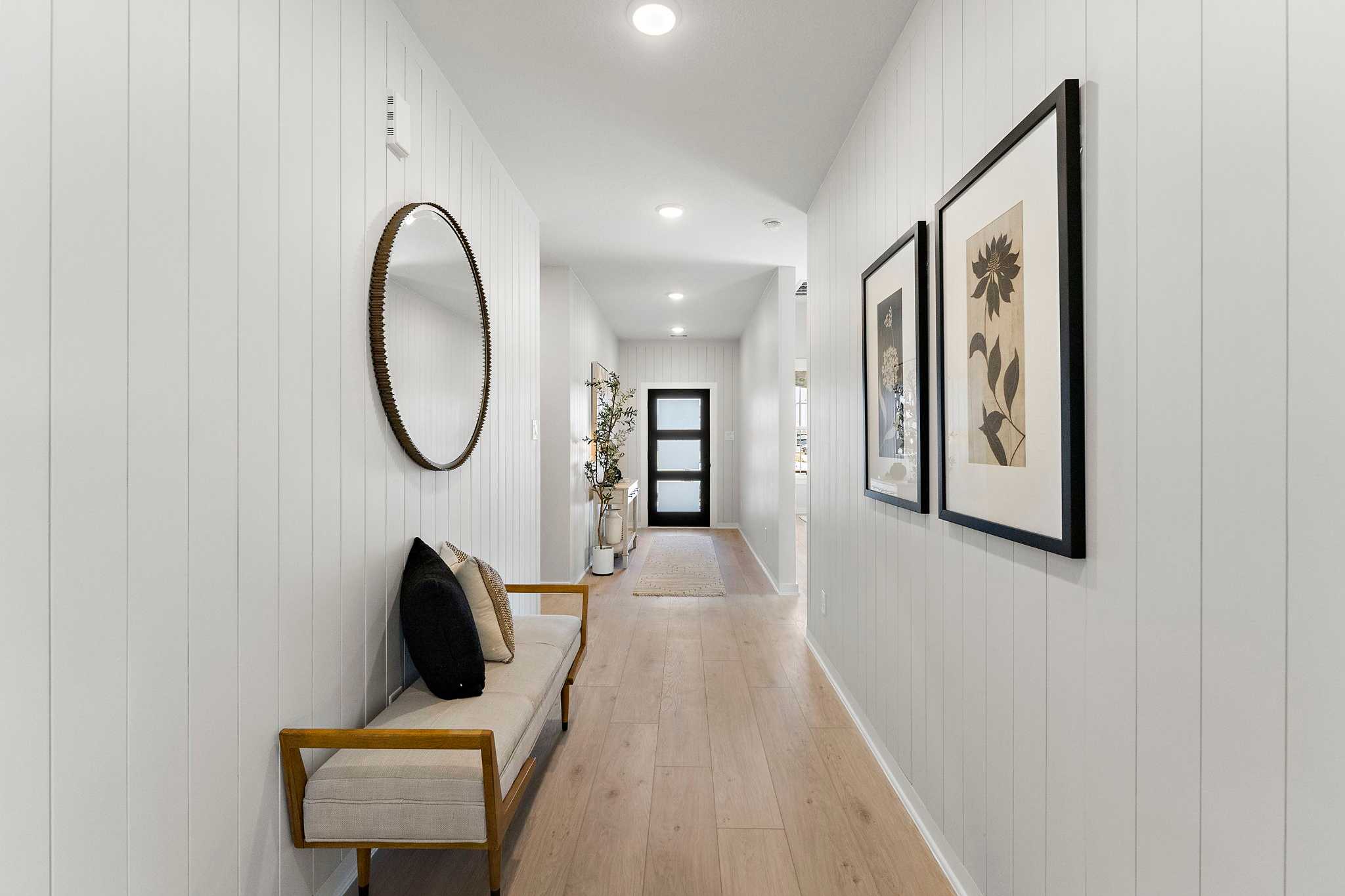 Bright hallway at Sundance Cove in Crosby, Texas with white shiplap walls, oak hardwood floors, bench, round mirror, and botanical art