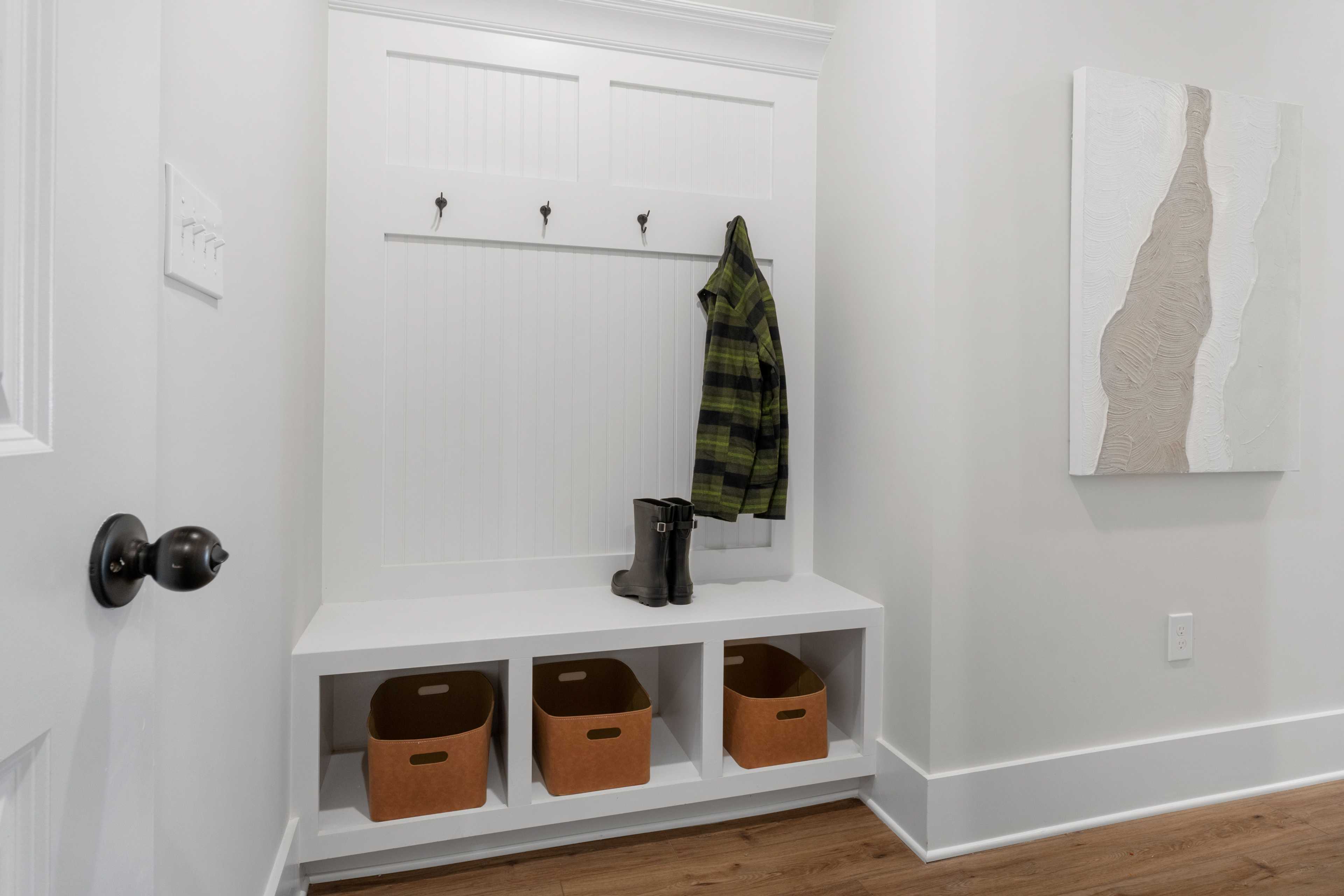 Spacious mudroom in Briercreek by Davidson Homes, Meridianville AL with white built-in storage, plaid jacket on hooks, boots, orange baskets, abstract art, hardwood floors