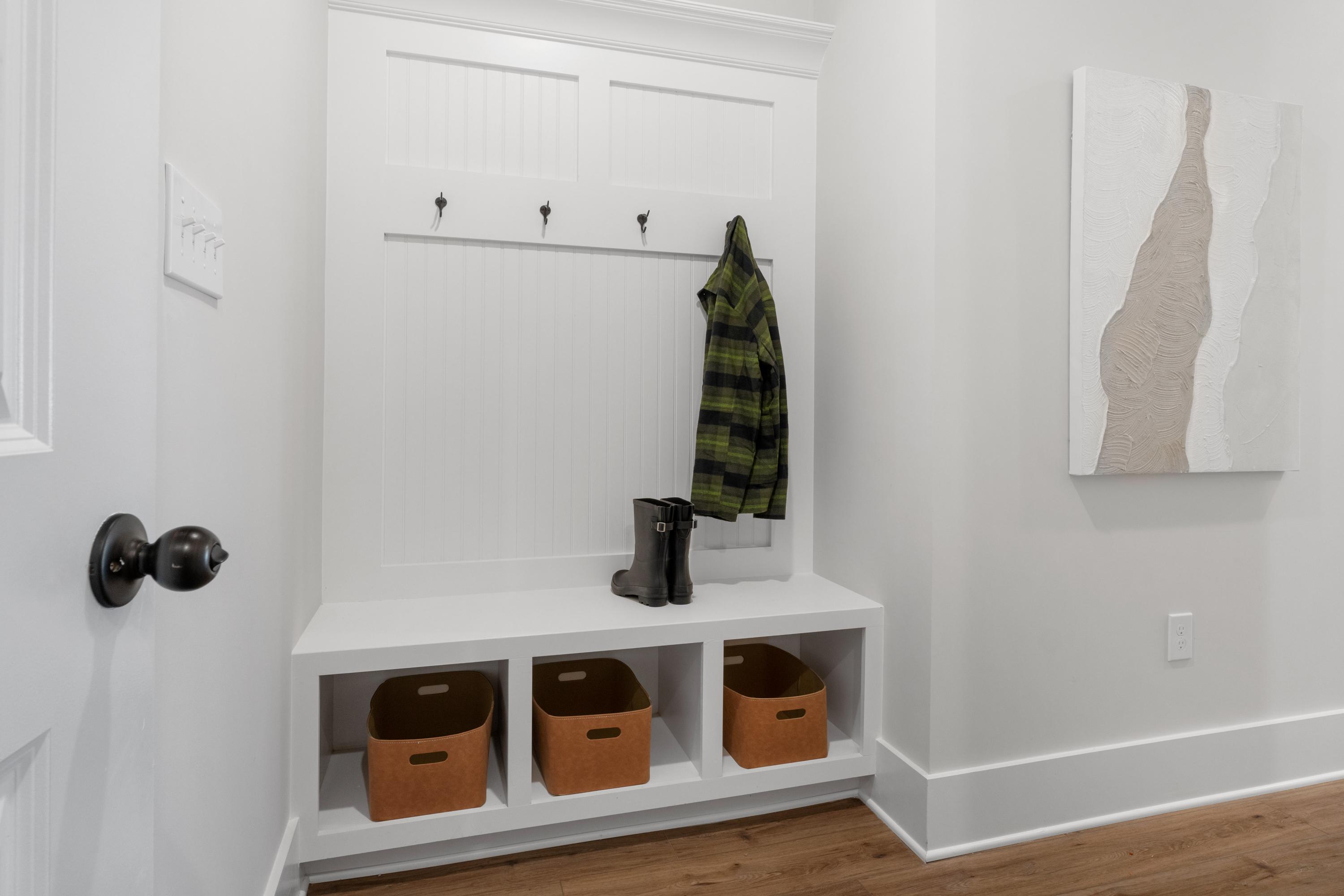 Spacious mudroom in Briercreek by Davidson Homes, Meridianville AL with white built-in storage, plaid jacket on hooks, boots, orange baskets, abstract art, hardwood floors