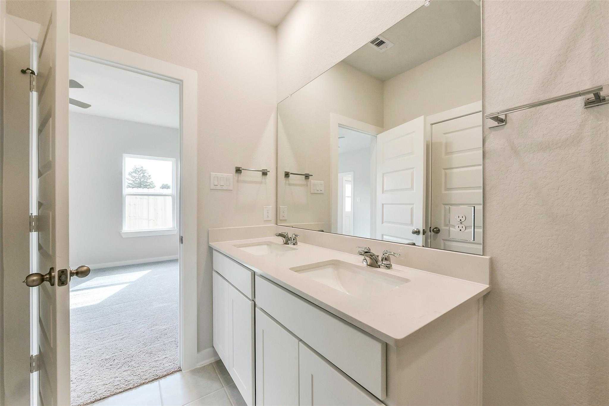 Modern master bathroom with double vanity sinks, large mirrors, and open bedroom door in Davidson Homes The Comal G, Dayton, Texas