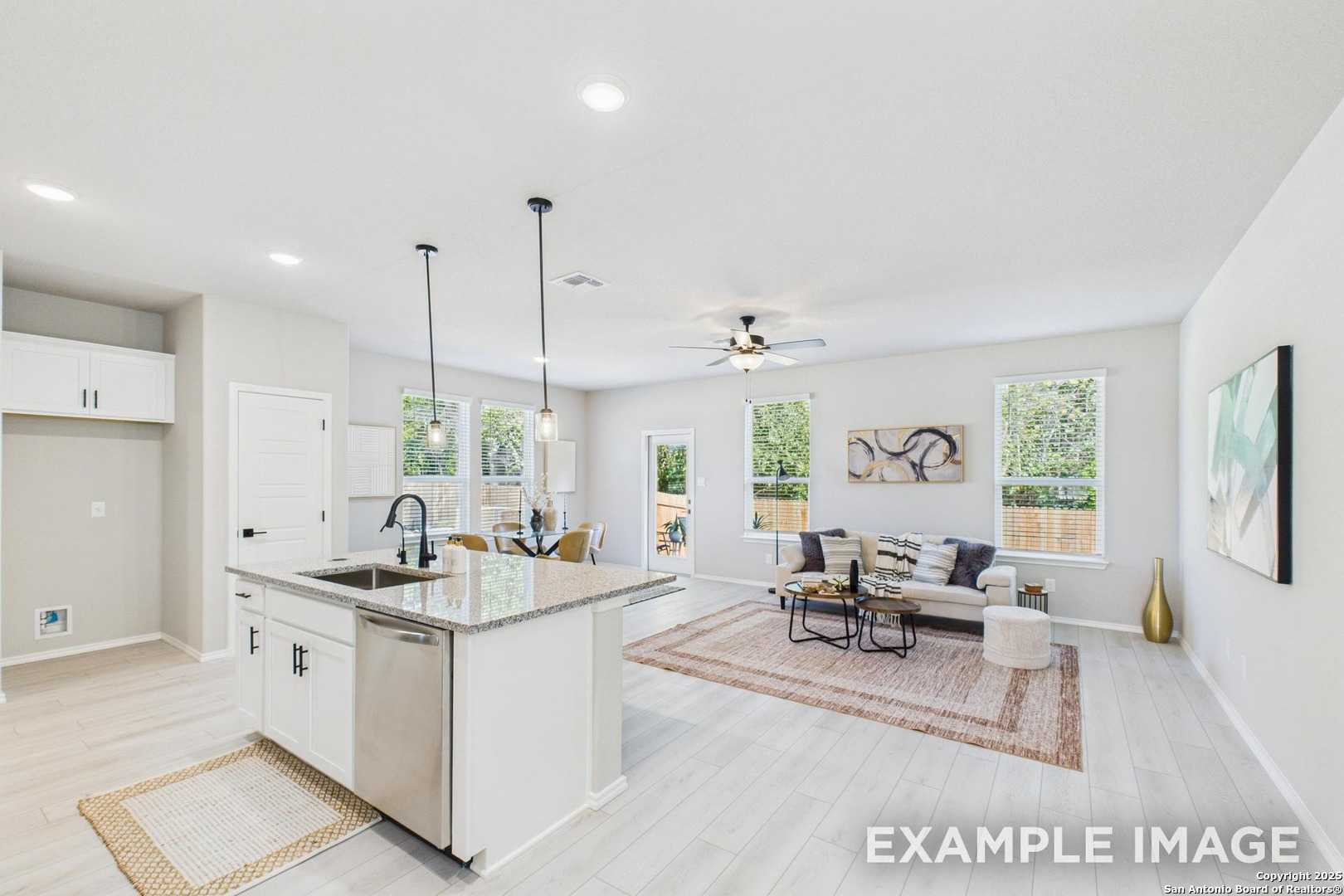 Modern open-concept kitchen with white island, stainless dishwasher, and sink adjoining living room with ceiling fan and sofa in The Charlotte B, San Antonio