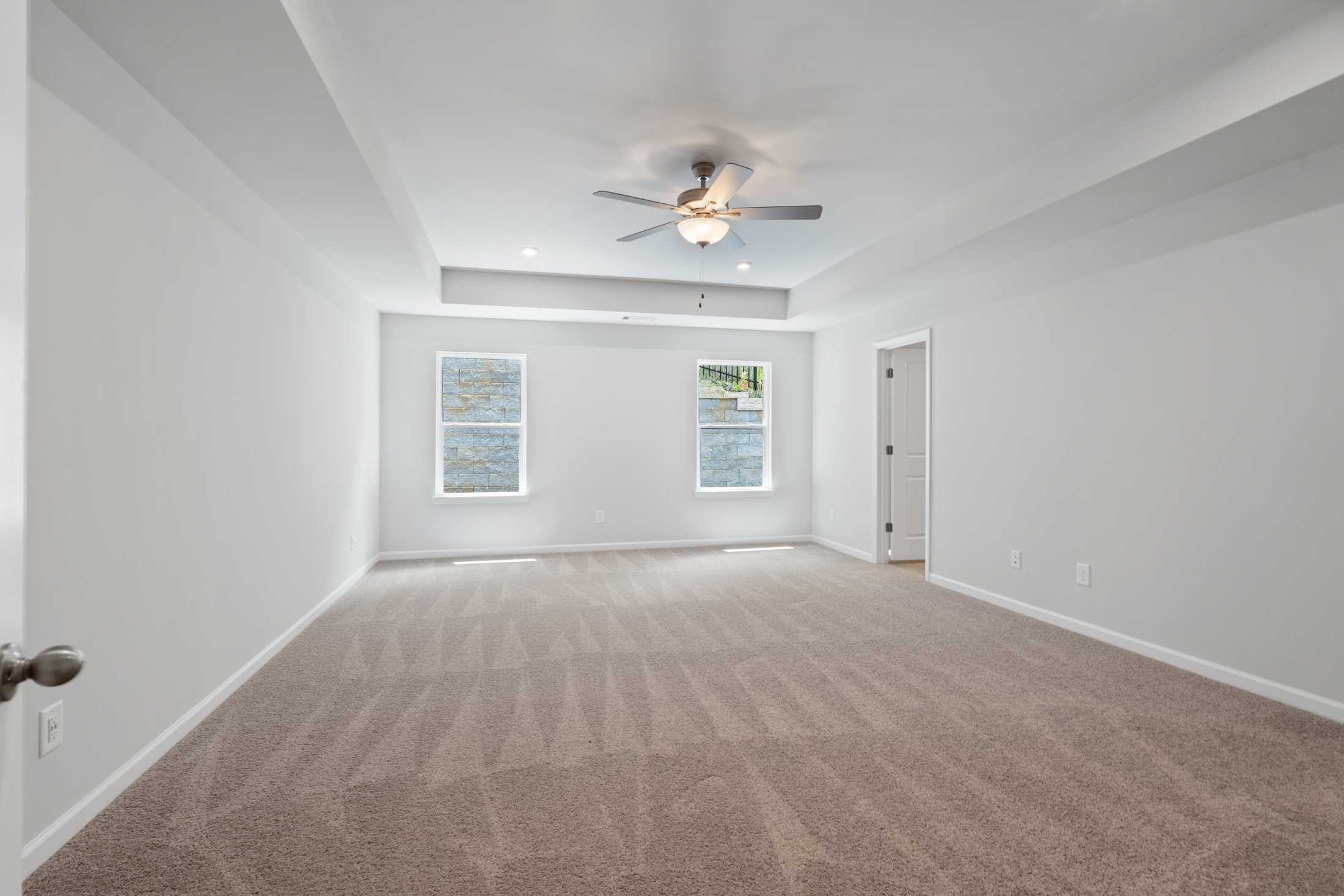 Spacious empty master bedroom in The Cary A with beige carpet, white walls, two large windows, and ceiling fan