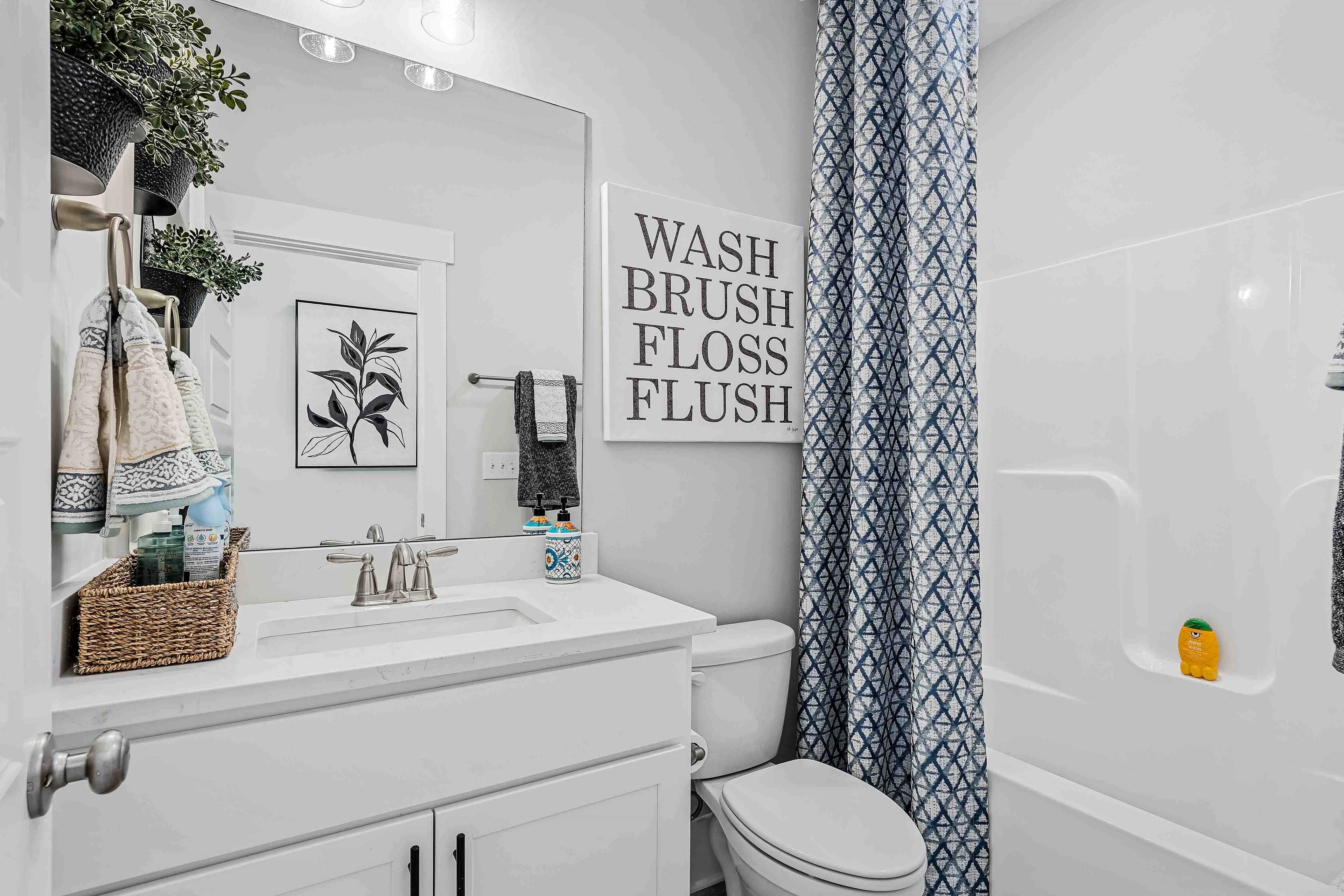 Cozy bathroom in Cain Park Hartselle AL with white vanity sink, blue shower curtain, toilet, tub, and "Wash Brush Floss Flush" sign