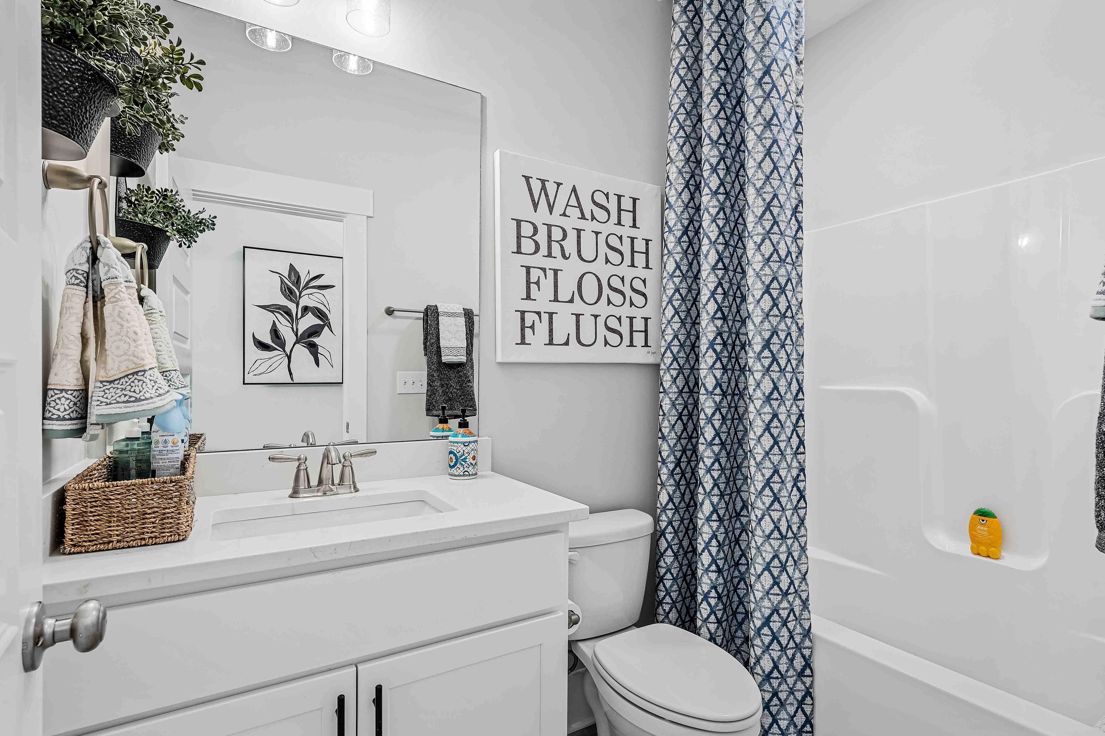 Cozy bathroom in Cain Park Hartselle AL with white vanity sink, blue shower curtain, toilet, tub, and "Wash Brush Floss Flush" sign