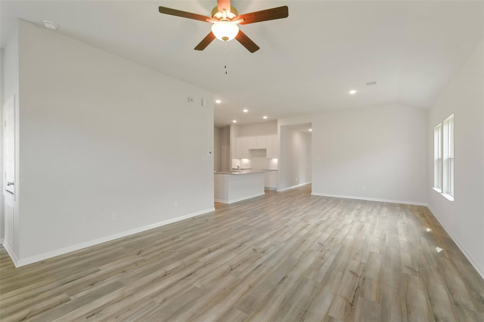 Open-concept living room with light wood floors, ceiling fan, and white kitchen view in Davidson Homes The Laguna B, Dayton Texas