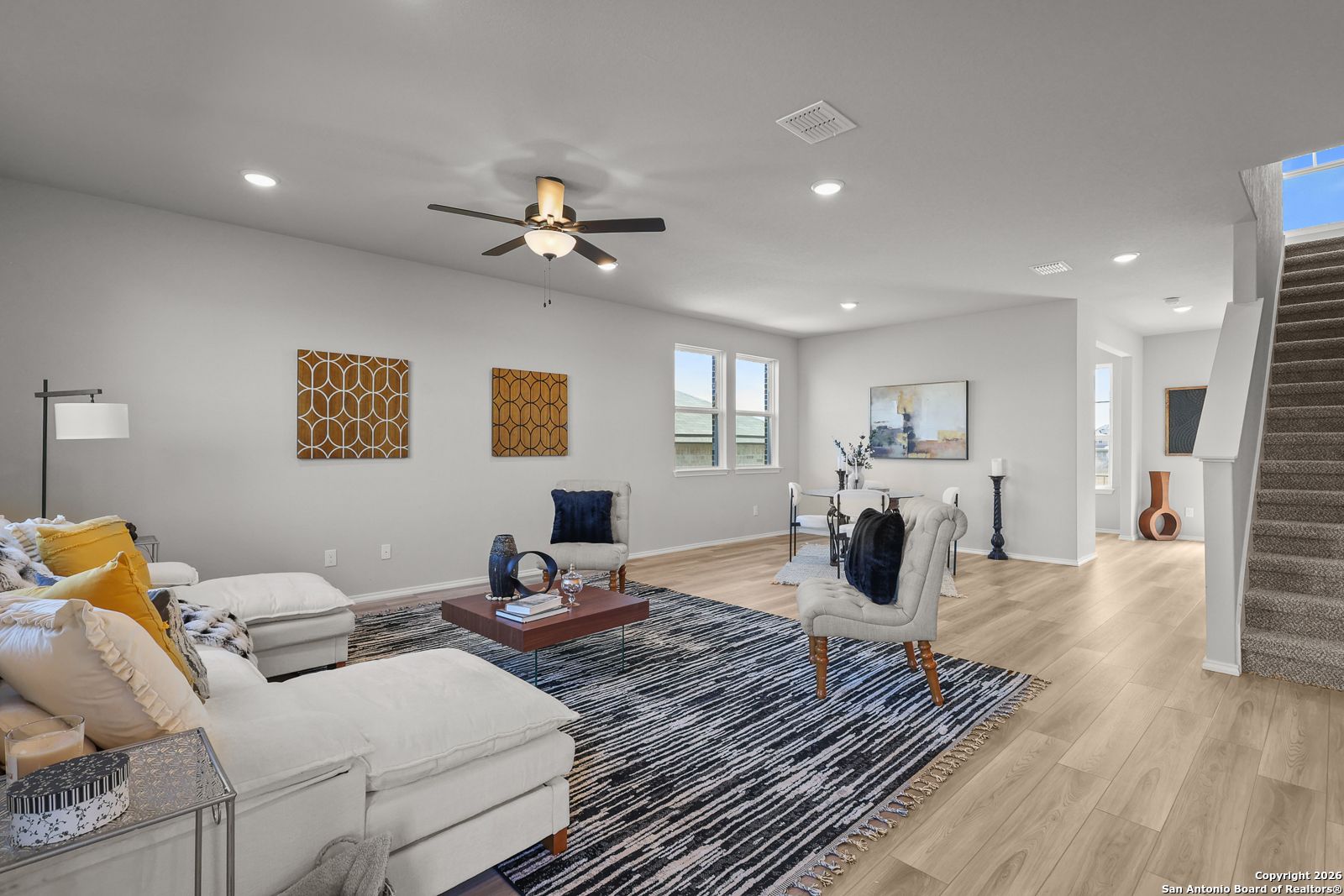 Modern living room with white sectional sofa, ceiling fan, open staircase, and artwork in Davidson Homes The Murray K, San Antonio, Texas