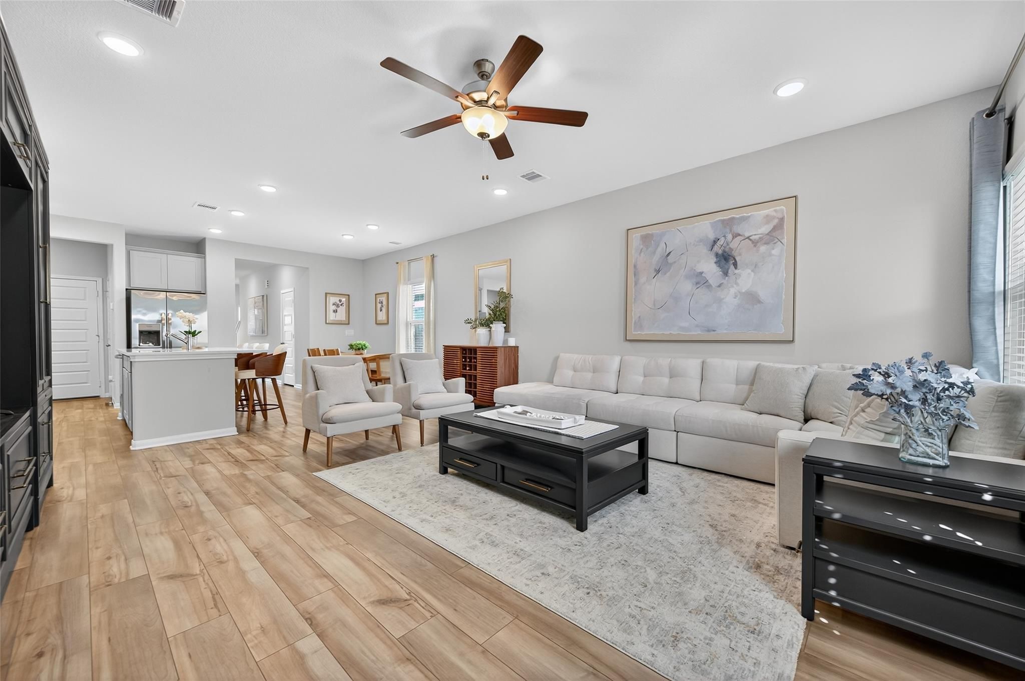 Open-concept living room with gray L-shaped sofa, ceiling fan, abstract art, and white kitchen island in Davidson Homes The Brazos E, Magnolia, Texas