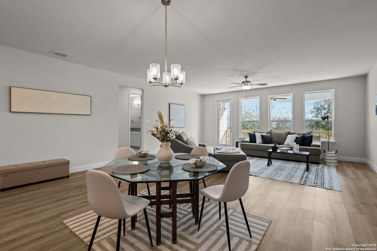 Open-concept dining-living area with glass table, gray sofa, large windows, hardwood floors in The Lanier H, Ladera, San Antonio