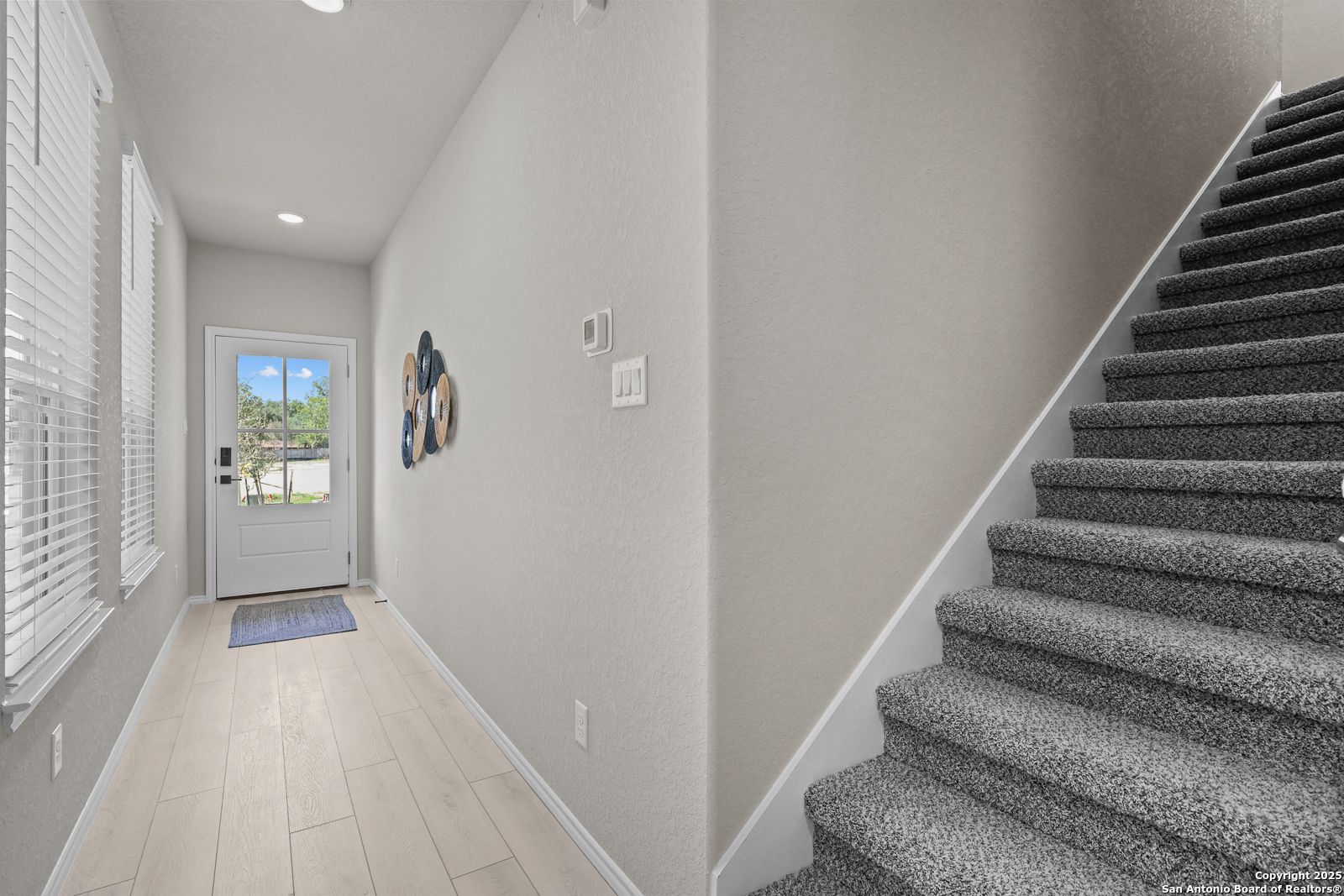 Bright entry foyer with light wood floors, neutral walls, carpeted staircase, and front door in Davidson Homes The Charlotte B, San Antonio