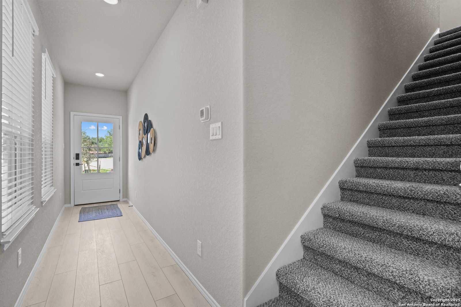 Bright entry foyer with light wood floors, neutral walls, carpeted staircase, and front door in Davidson Homes The Charlotte B, San Antonio
