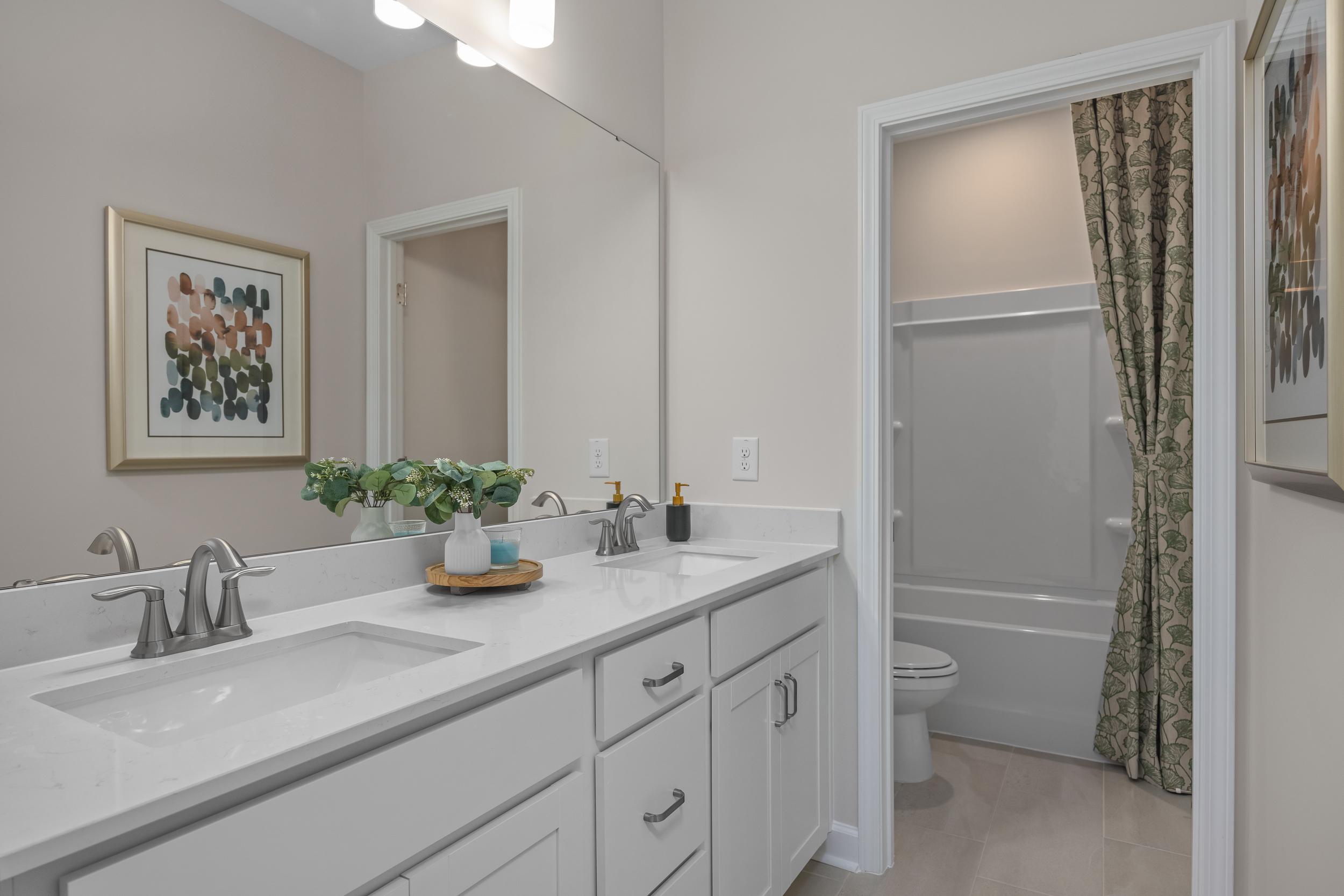 Spacious master bathroom in The Hickory II with double white vanity, large lighted mirror, potted plants, and curtained tub-shower