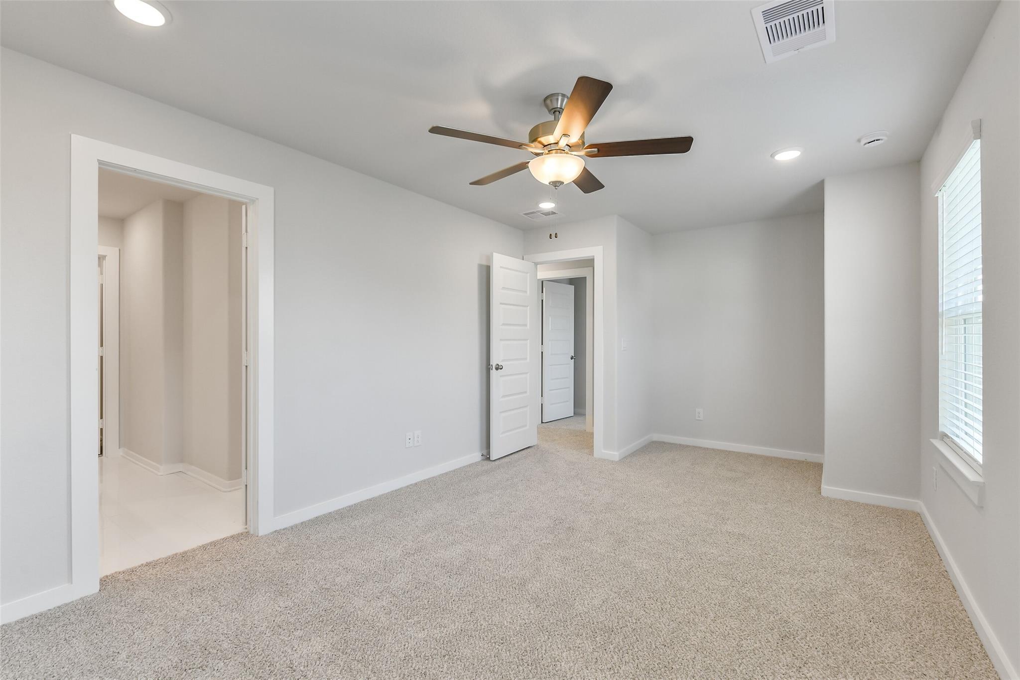 Spacious bedroom with light gray walls, beige carpet, ceiling fan, and en-suite bath door in Davidson Homes The Trinity F, Magnolia, Texas