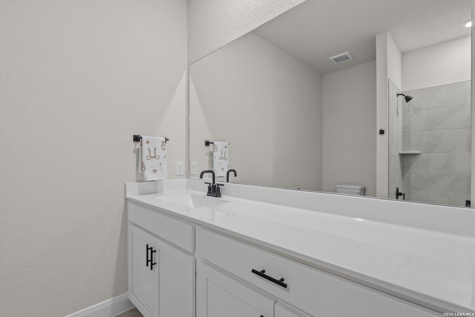 Modern bathroom with white double vanity, quartz countertop, large mirror, and tiled shower in Davidson Homes The Lanier G, Castroville TX
