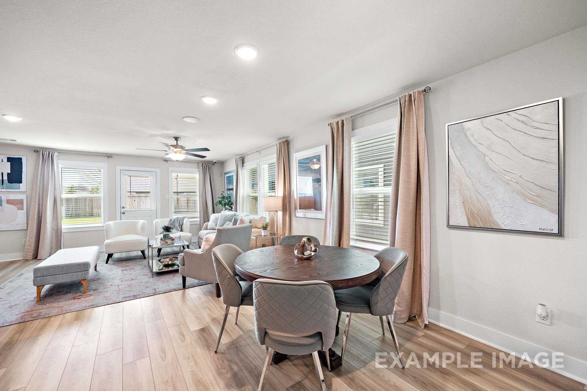 Open-concept living and dining space in The Lily A featuring beige sofas, round wooden table, ceiling fan, abstract wall art, and hardwood floors