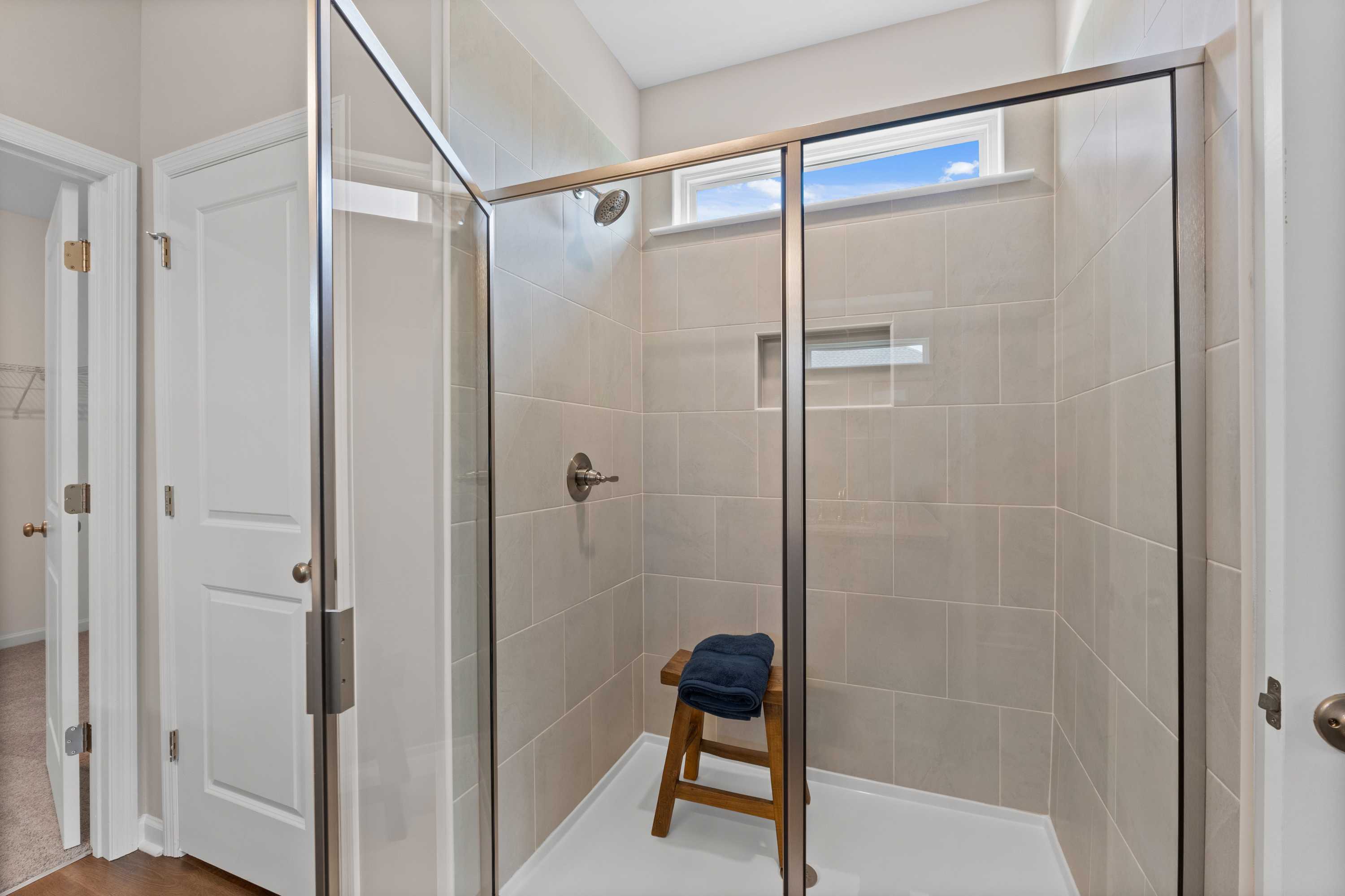 Spacious walk-in shower at Riverton Preserve in Huntsville AL featuring glass enclosure, beige tile walls, wooden bench, and niche shelf