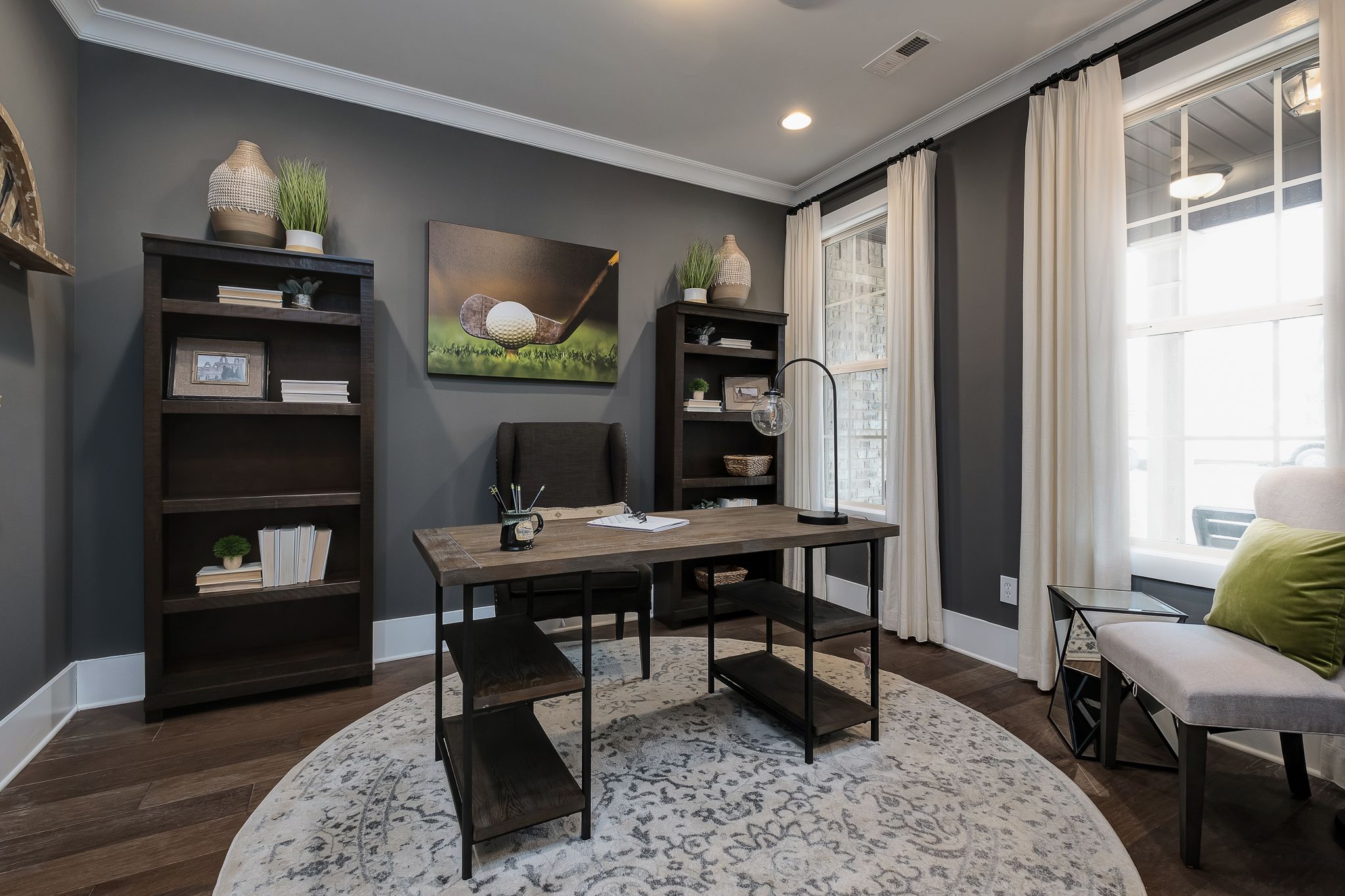 Cozy home office in Chimney Creek, Hampton Cove AL with rustic wooden desk, bookshelves, golf art, gray walls, and large windows by Davidson Homes