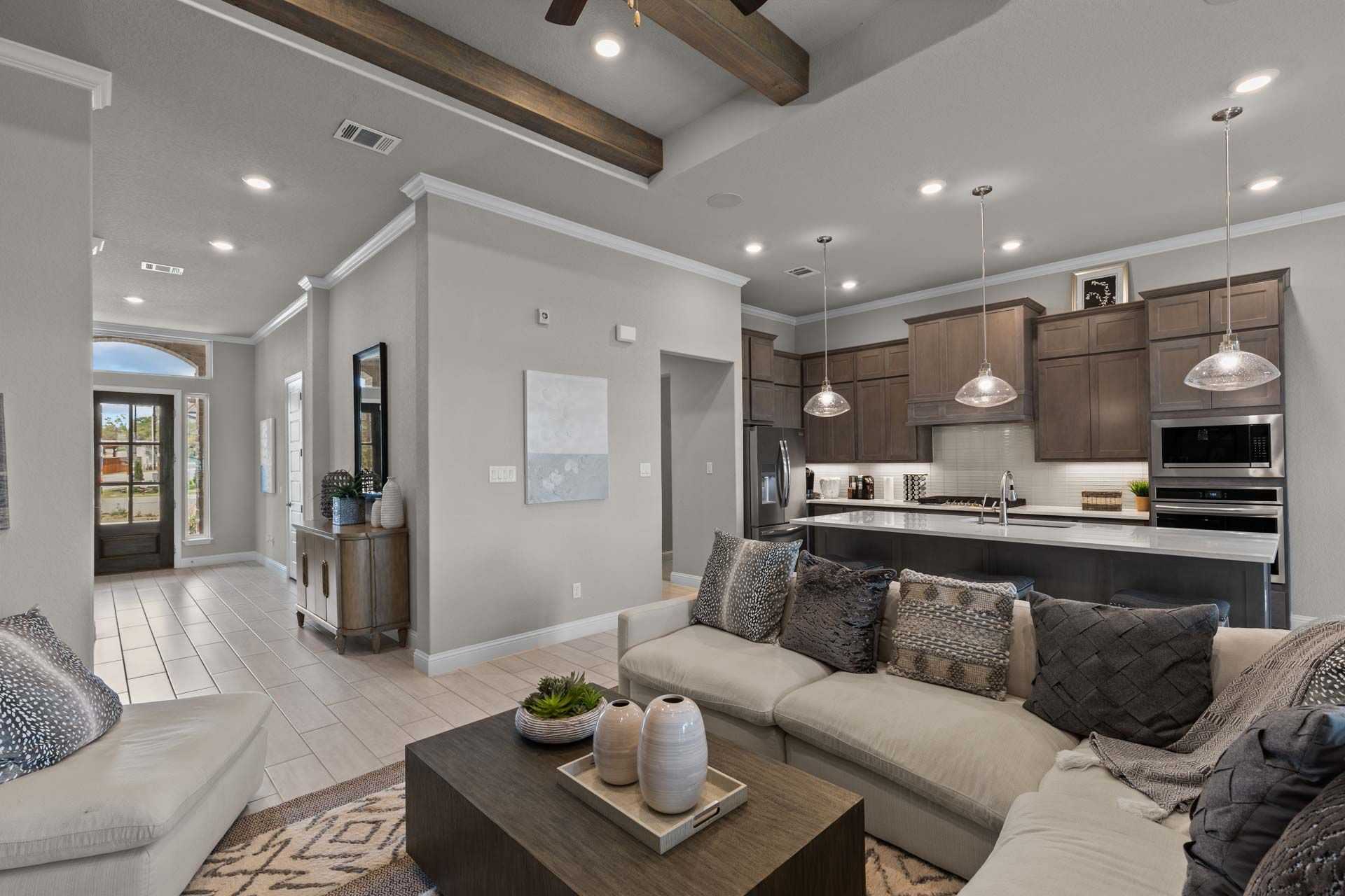 Open-concept living room and kitchen in Ladera San Antonio TX home by Davidson Homes featuring gray walls, wooden beams, sectional sofa, and modern island