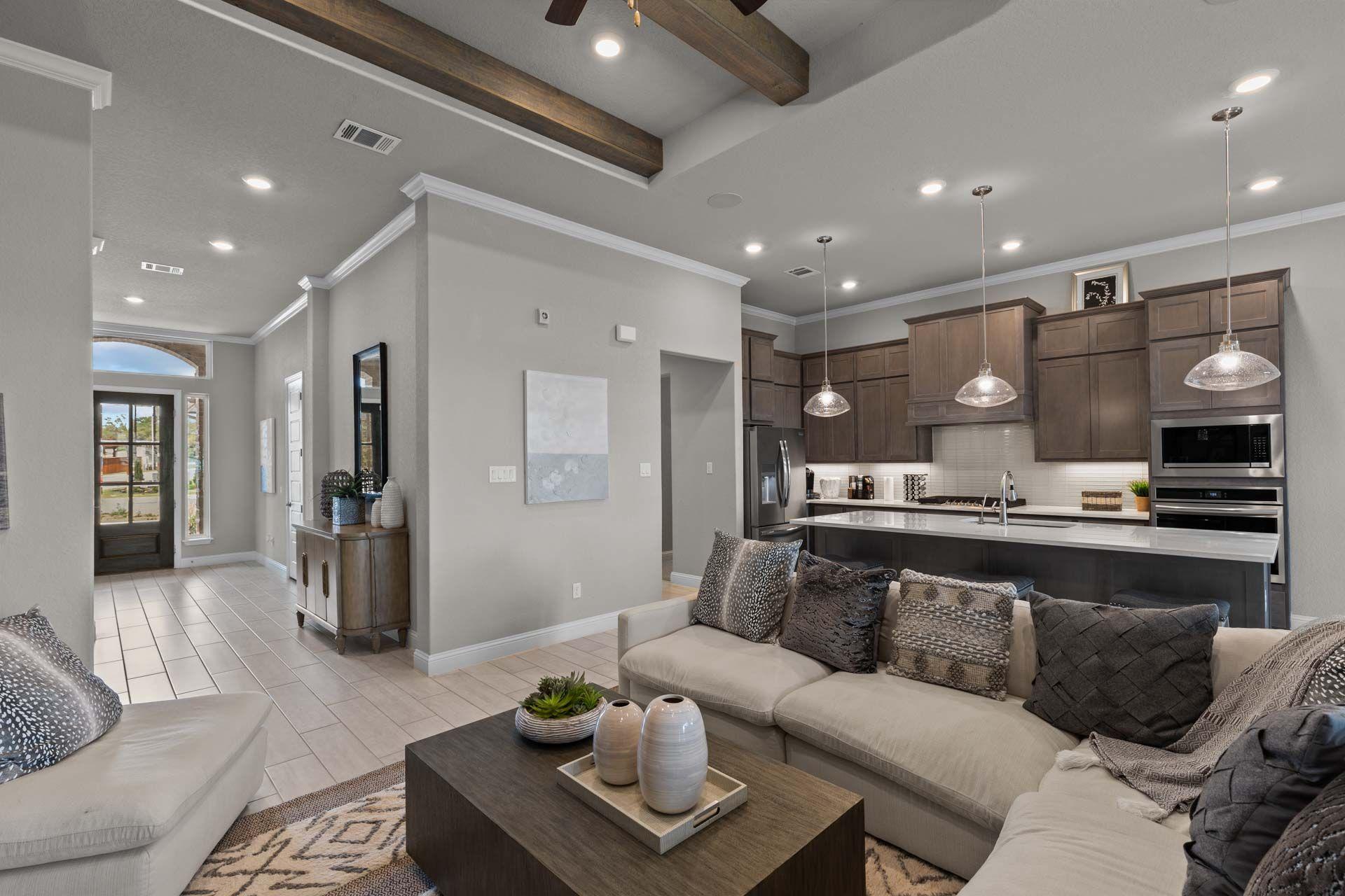 Open-concept living room and kitchen in Ladera San Antonio TX home by Davidson Homes featuring gray walls, wooden beams, sectional sofa, and modern island
