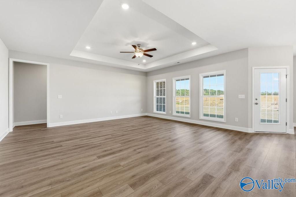 Spacious empty living room with tray ceiling, ceiling fan, large windows, and hardwood floors in The Harrison floor plan, Hartselle, Alabama