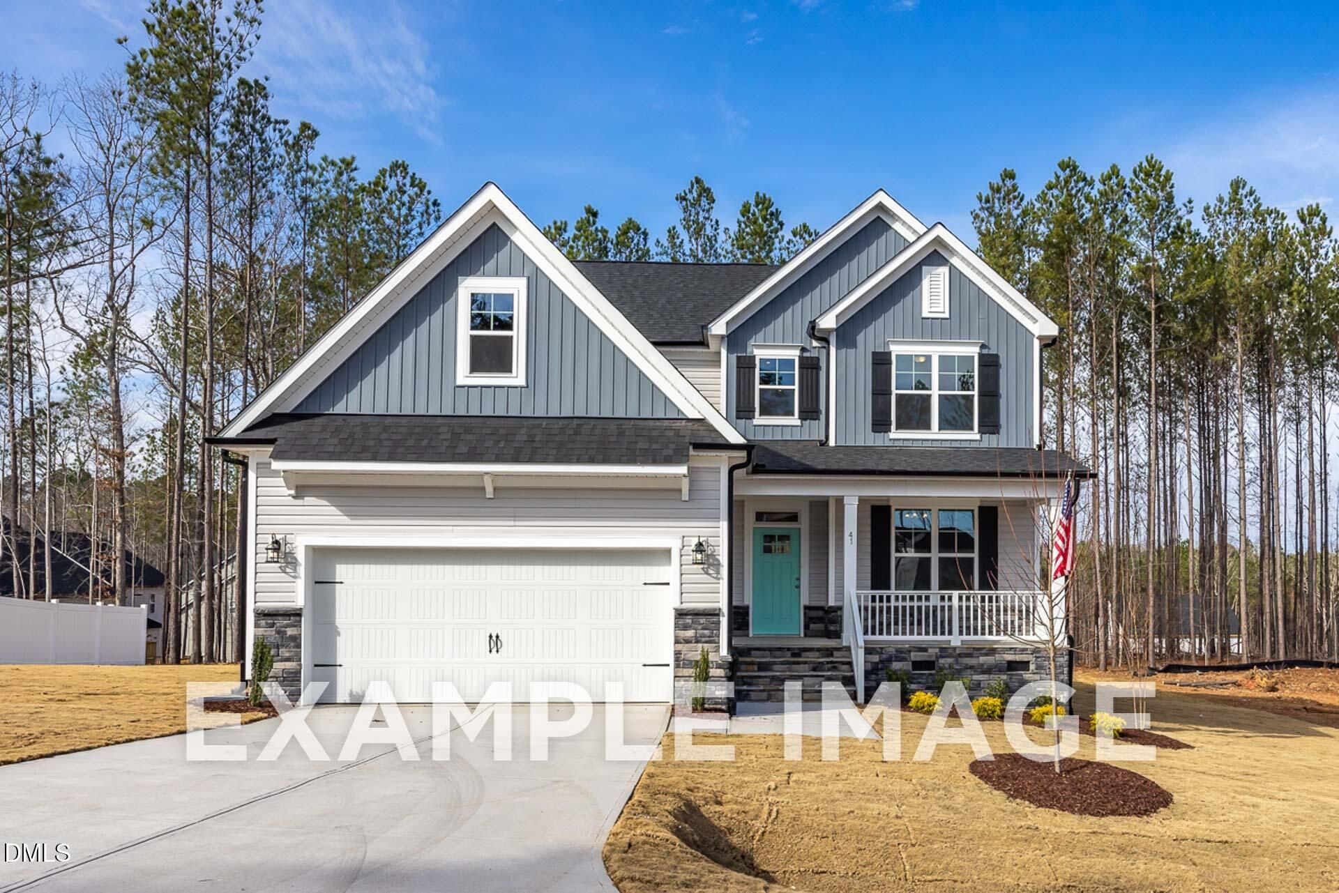 Two-story blue Davidson Homes The Ash B with 2-car garage, front porch, and American flag amid pines in Woodland Crossing, Zebulon, NC