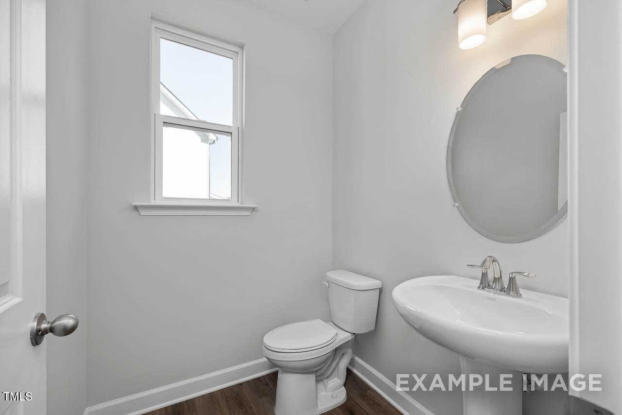 Bright powder room with round mirror, pedestal sink, toilet, and window in Davidson Homes The Hickory II B, Lillington, NC