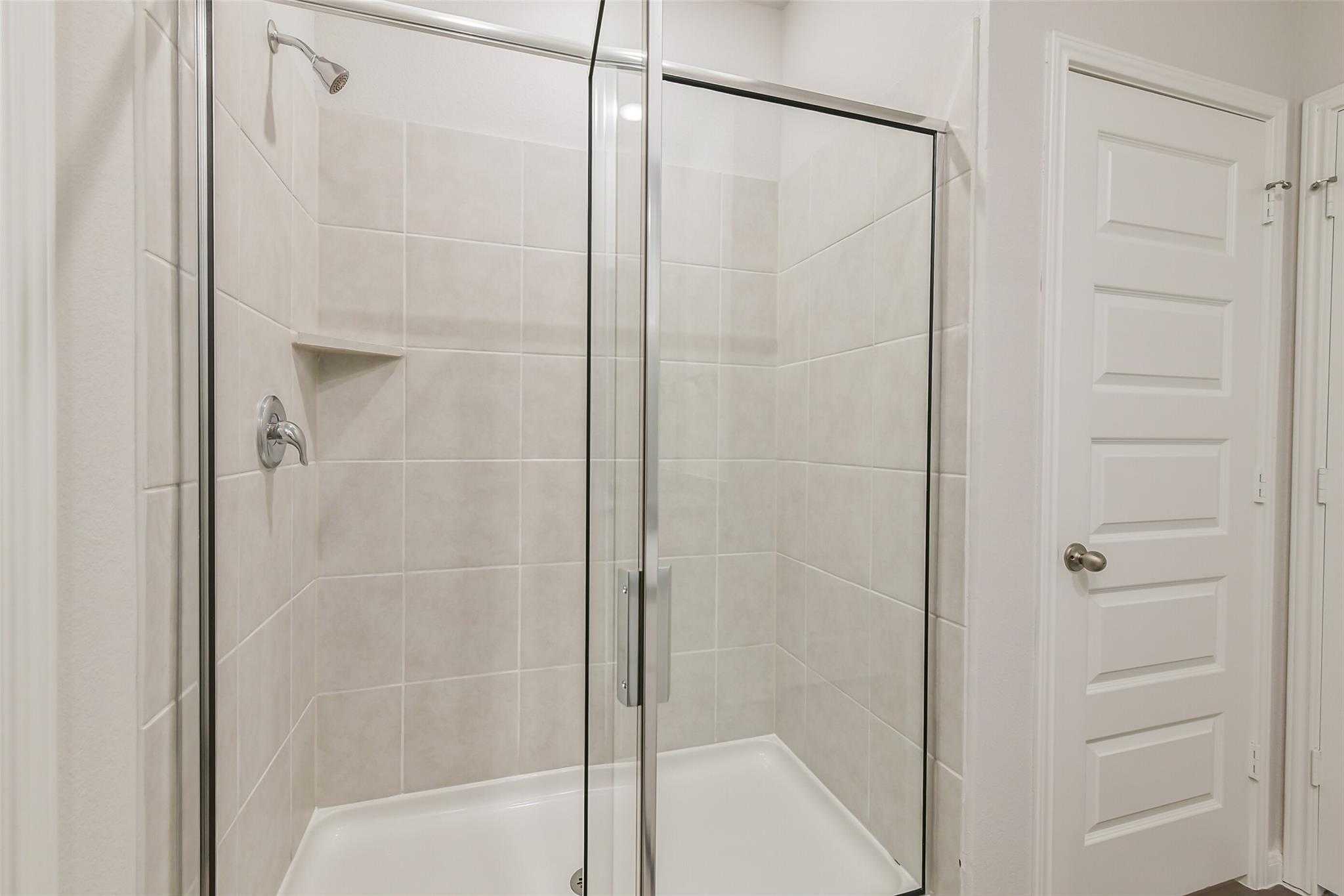 Modern walk-in shower with frameless glass enclosure, beige subway tiles, and built-in shelf in Davidson Homes The Brazos F, Conroe, Texas